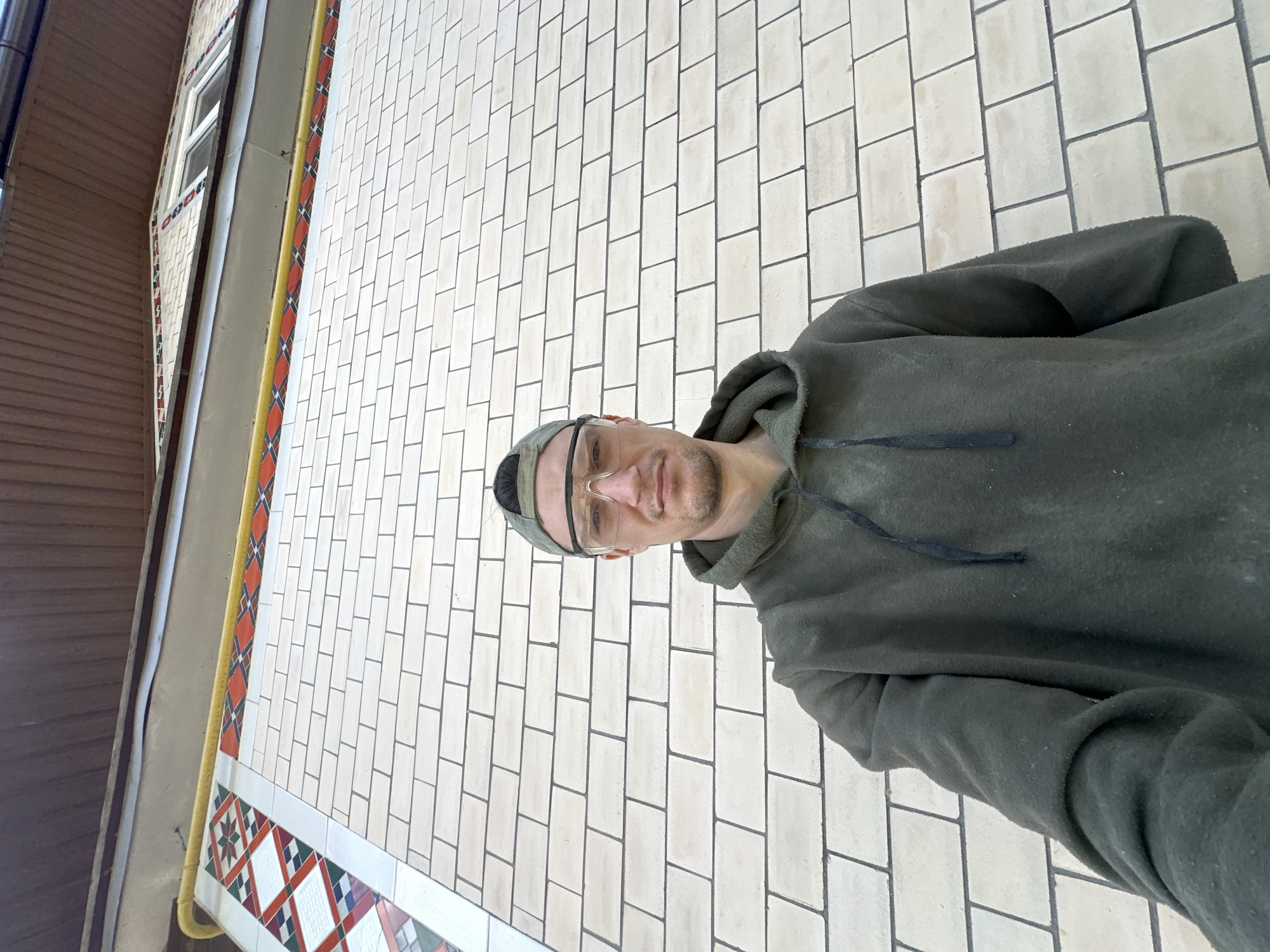 A low-angle, wide shot shows a man standing in front of a light-colored brick wall. He is wearing a dark green hoodie, a green baseball cap, and clear safety glasses. The man is facing the camera and appears to be taking a selfie. The brick wall is laid in a running bond pattern with dark gray mortar. Above the brick wall, there is a decorative band of tiles with a geometric pattern in red, white, blue, and green, bordered by a yellow pipe. The roofline of a building is visible at the top of the frame, with brown ribbed siding. A small section of a window is visible to the right, high up on the building. The lighting suggests it is daytime, possibly overcast. The overall scene suggests an outdoor construction or renovation site. The location is identified as Makiv, Ukraine.