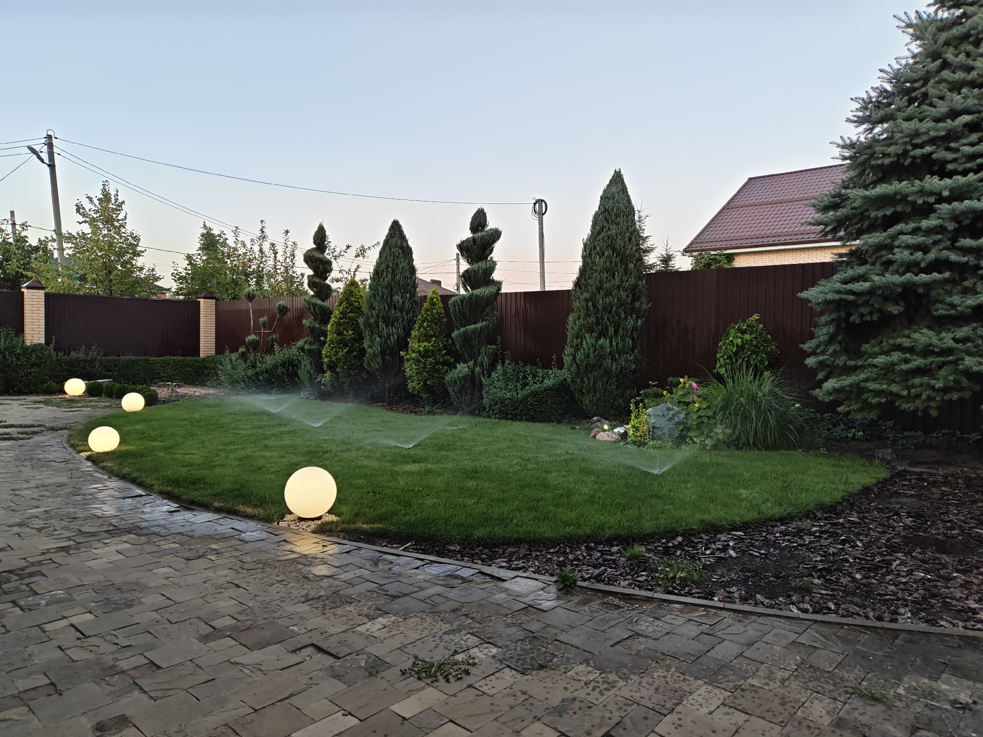 The image depicts a manicured garden scene at dusk in Vodopadnyy, Russia. A curved, well-maintained lawn is being watered by sprinklers, with visible arcs of water misting the grass. The lawn is bordered by a dark brown wooden fence and a variety of evergreen shrubs and trees, including several topiaries. Illuminated by soft, warm light are four large, spherical outdoor lamps placed strategically along a winding paved pathway. The pathway is constructed from irregularly shaped, grey paving stones, which appear slightly wet. In the background, the roofline of a house is visible above the fence, and utility poles with power lines stretch across the pale sky. A large coniferous tree stands prominently on the right side of the frame. The overall impression is one of a carefully tended, modern outdoor living space at the transition from day to night.