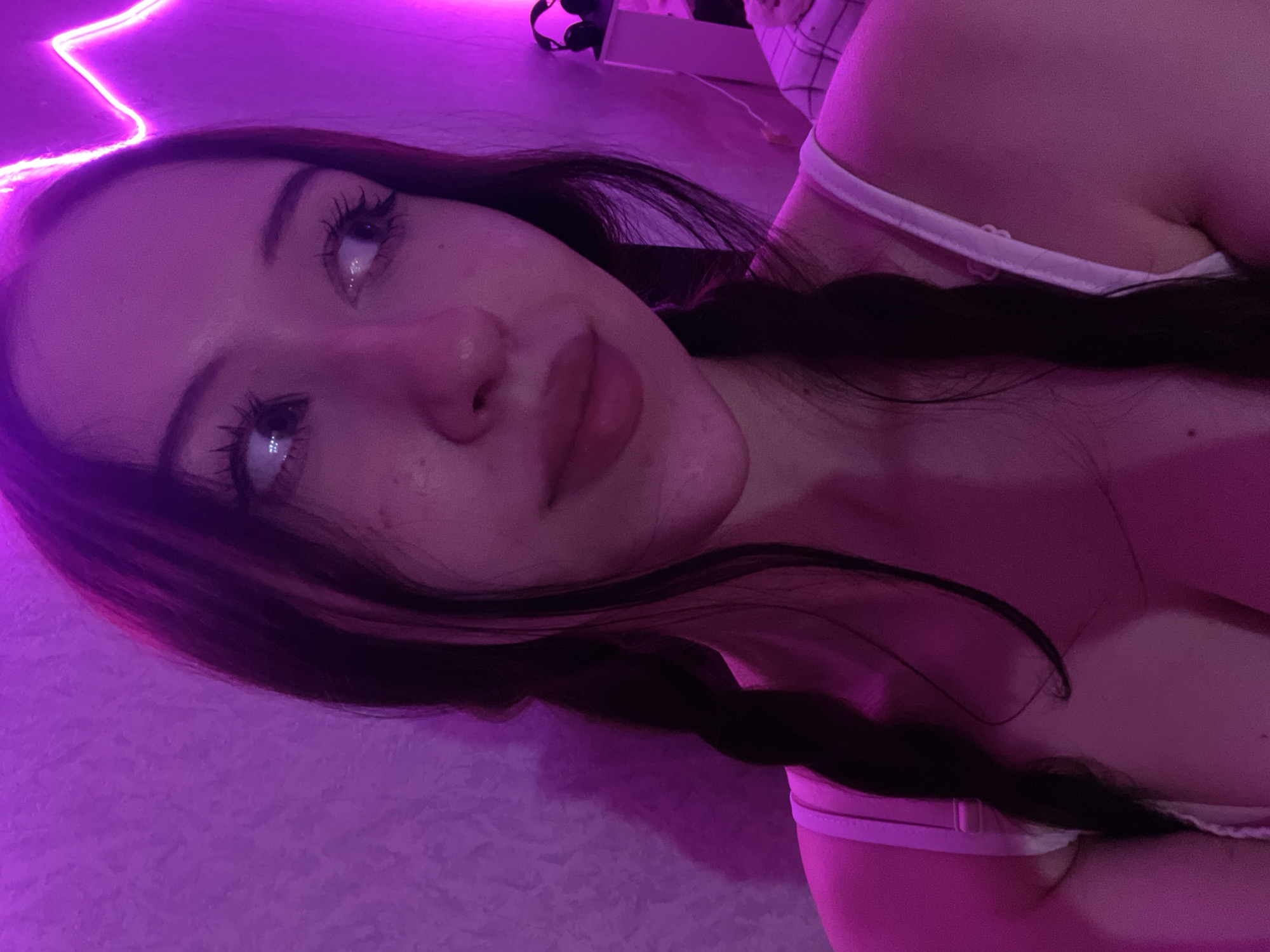 A young adult female is depicted in a close-up, self-portrait style photograph, illuminated by strong magenta and purple neon lighting. She has dark hair styled in two distinct braids, which are visible resting on her shoulders and chest. Her eyes are looking upwards and to her right, with visible eyelashes and subtle freckles on her cheeks. Her lips appear full and slightly glossy, with a soft, candid expression. She is wearing a white garment with visible straps, likely a bra or camisole. The background features a textured wall, with a zigzag-shaped neon light strip mounted on the upper right side. To the far right, a pair of dark headphones rests on a small, white piece of furniture, with a checkered textile (possibly bedding) partially visible beneath it. The specific location in the city of Votkinsk, Russia, cannot be determined from the visual information provided in the image.