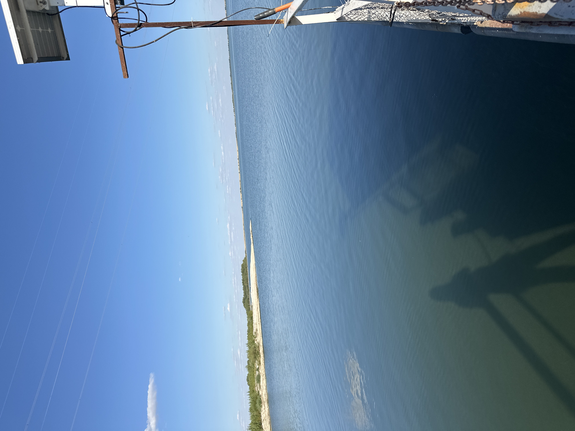 The image depicts a serene body of water, likely a bay or lake, under a clear blue sky with scattered white clouds. In the distance, a sandy shoreline is visible, fringed with green vegetation and trees. The foreground features a body of water with visible ripples and a shadowy silhouette of a person or people on the right side of the frame, cast by an unseen structure or object. To the far right, part of a metallic structure with a solar panel on top is visible, along with chains and cables. The scene suggests a tranquil outdoor setting, potentially a waterfront area or a pier. Without additional context, it is not possible to confirm the specific location within the city of Приморск, Russia.