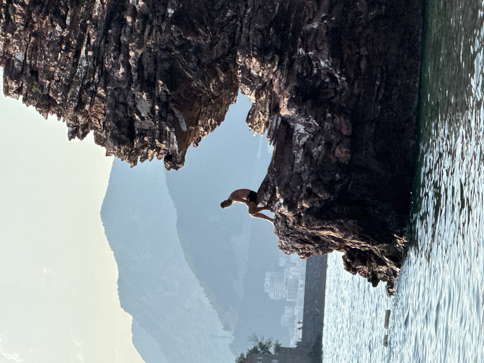 A person is captured mid-leap, jumping from a rocky outcrop into the sea. The scene is set against a backdrop of mountains and a hazy sky, suggesting a warm, possibly early morning or late afternoon. The water is calm and reflects the sunlight, creating a shimmering effect. The rugged texture of the rocks contrasts with the smooth surface of the water and the soft, muted tones of the distant landscape. The orientation of the image is such that the viewer perceives the scene as if upside down. The image was taken in Sveti Stefan, Montenegro.