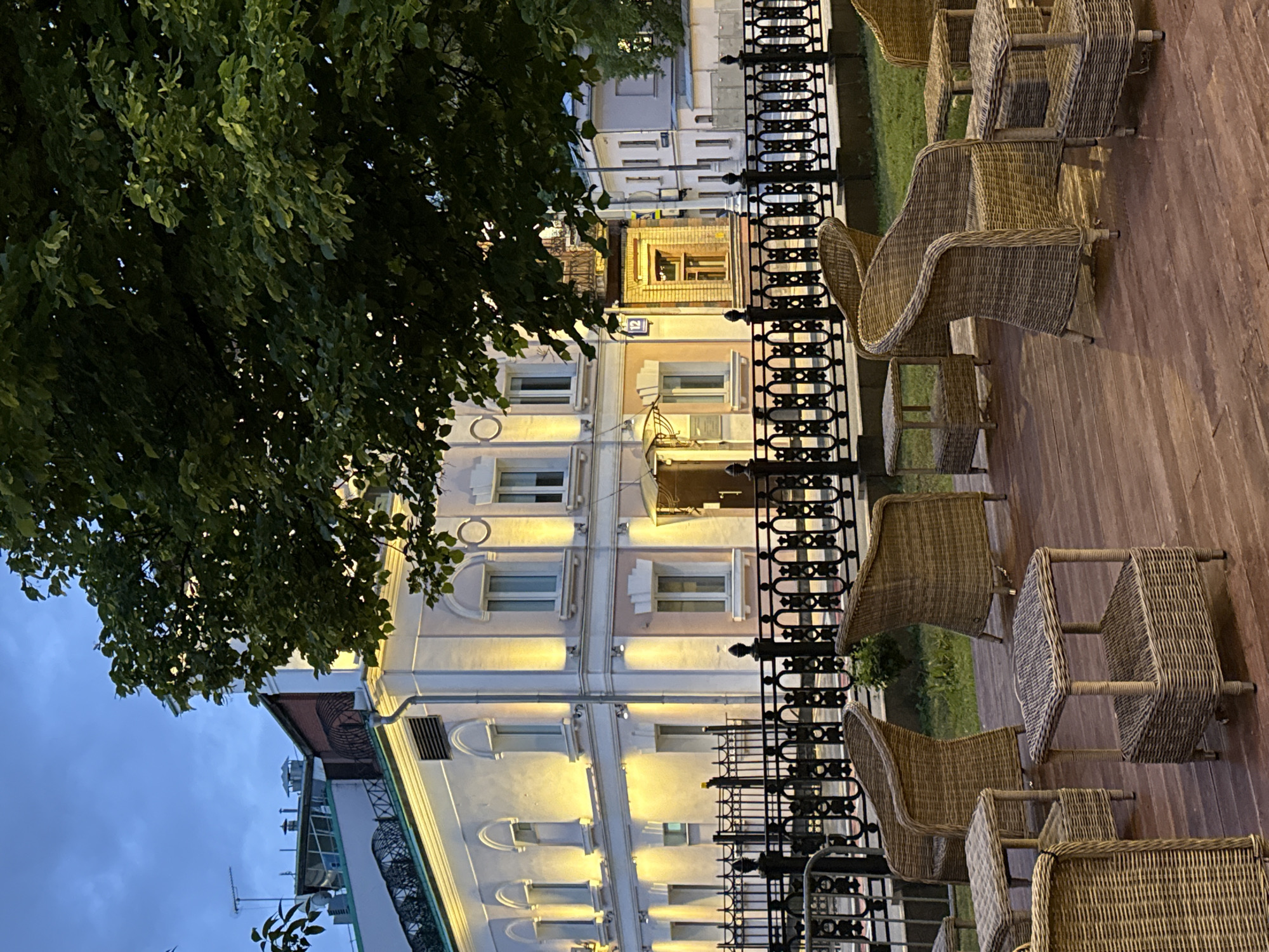 The image shows an outdoor cafe in Moscow, Russia. The cafe has wicker chairs and tables arranged on a wooden platform. A decorative black metal fence separates the cafe from the street. Behind the fence, there is a multi-story building with illuminated windows. A tree with dense foliage is visible on the left side of the image. The sky is a dusky blue.