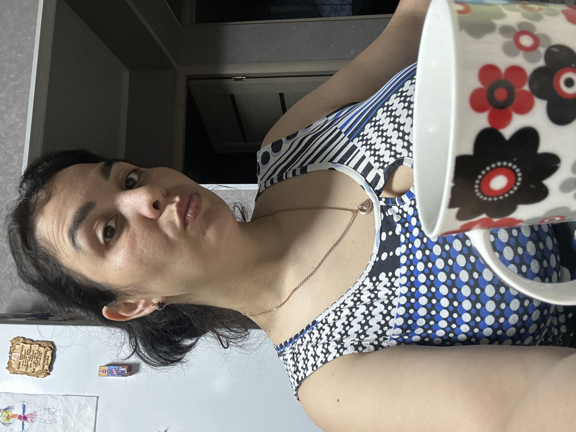 The image captures a woman in a close-up, rotated perspective, appearing as if she is lying down or leaning back while taking a selfie. Her head is positioned on the left side of the frame, and she looks directly at the camera with a playful, slightly pouting expression. She has dark hair and eyes and wears a sleeveless top patterned with blue, white, and black geometric shapes and polka dots. Around her neck is a thin gold chain necklace with a small pendant, and a small earring is visible in her right ear.
A white mug, decorated with bold red, black, and grey floral patterns, is held or positioned in front of her right side, partially obscuring her. The background behind her left shoulder reveals a dark interior space with a door frame. To her right, a light-colored vertical surface, likely a refrigerator, is decorated with two magnets and a child's drawing. One magnet is shaped like an aged scroll and displays Cyrillic text, which includes phrases such as "ЧТО ПОЖЕЛАЕШЬ ТО И..." (What you wish, that...) and "ДОМА" (at home), suggesting a blessing or motivational message. The second magnet is a small, colorful rectangular box. The scene is lit by indoor lighting.