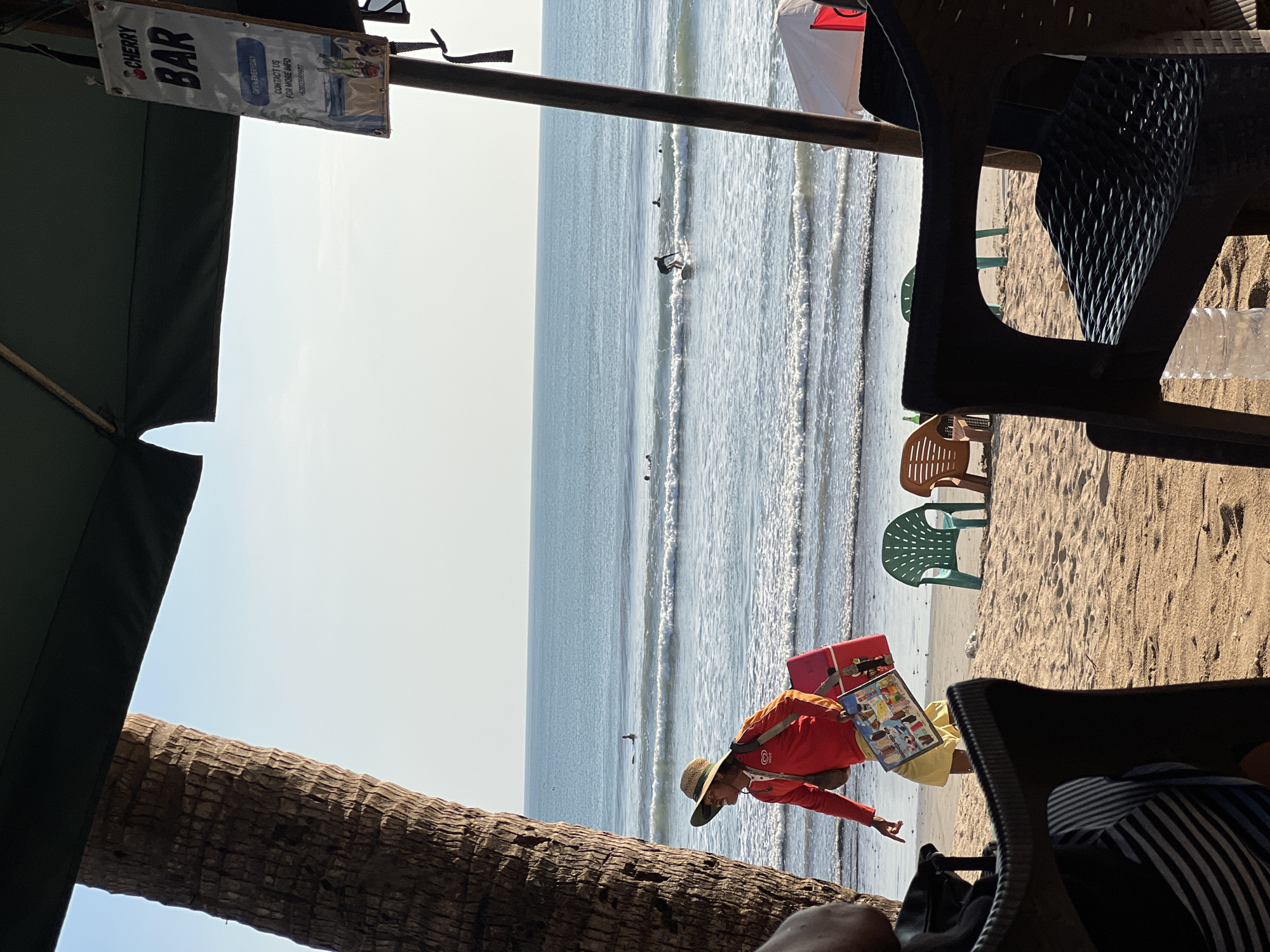 This image depicts a beach scene in Kuta, Indonesia, with a focus on a person walking on the sand near the water's edge. The person, wearing a red long-sleeved shirt, a straw hat, and yellow shorts, carries a pink cooler and a board displaying images of various items, possibly for sale. In the background, the ocean stretches to the horizon with several surfers visible in the water. The foreground shows beach chairs and a part of an umbrella, suggesting a seating area for beachgoers. A sign for "Cherry Bar" is attached to a pole, indicating the presence of a nearby establishment. The lighting suggests it is daytime, with clear skies and bright sunlight. The overall impression is one of a vibrant beach atmosphere in a popular tourist destination.