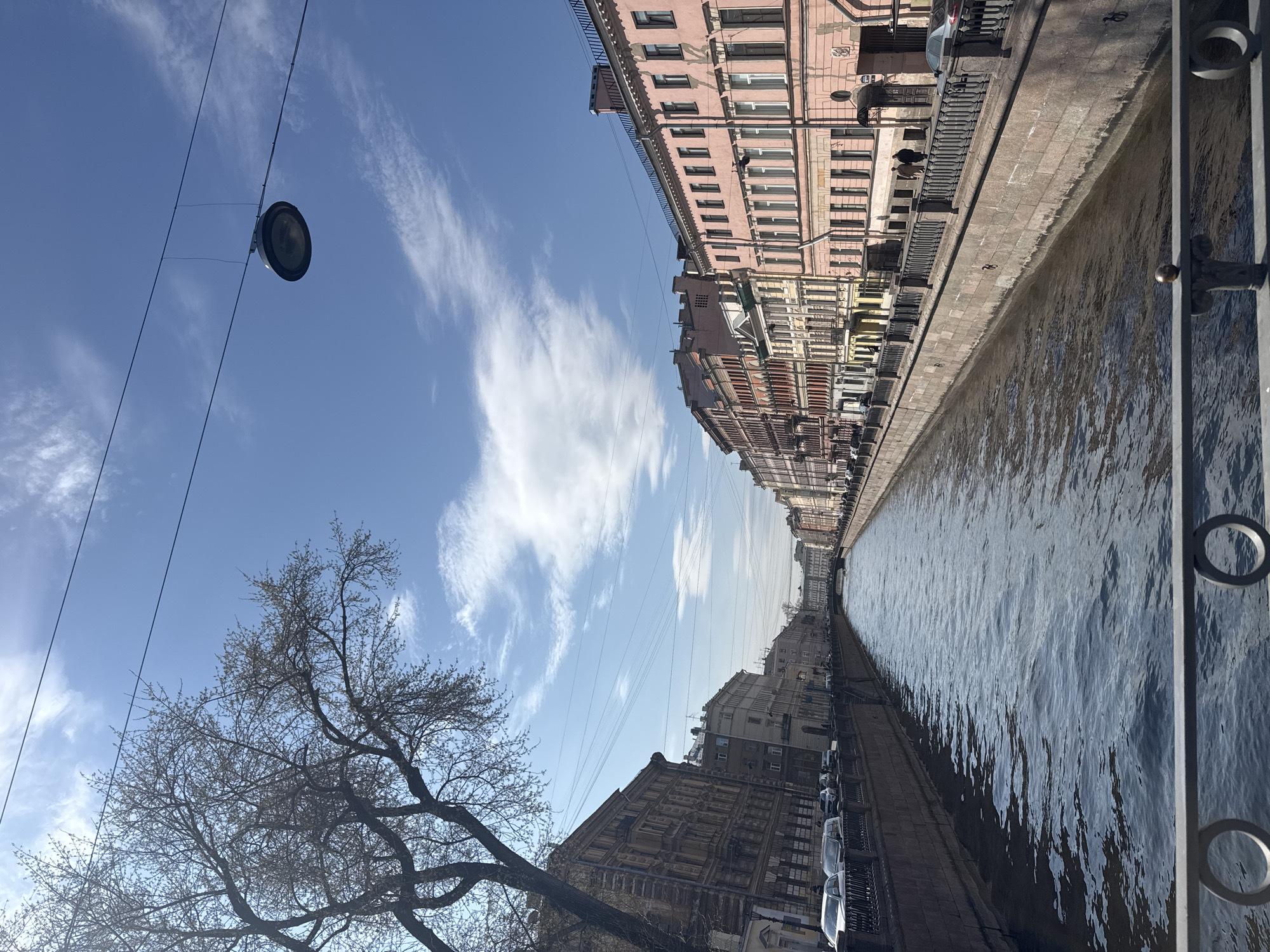 This image depicts a serene canal scene in Saint Petersburg, Russia, viewed from a slightly elevated position. The perspective looks down the length of the canal, which runs through an urban landscape under a bright, clear blue sky adorned with scattered wispy white clouds.
On the left side of the canal, a row of multi-story historic buildings with classical architecture lines the embankment, extending into the distance. Some buildings are painted a soft pink, while others exhibit darker, more weathered facades with numerous windows and architectural details. Cars are parked along the embankment in front of these buildings. In the lower-left foreground, a large deciduous tree, currently bare of leaves, its intricate branches reaching towards the sky, suggests the season is late winter or early spring. Overhead, several thin cables or power lines crisscross the sky, with a circular street lamp suspended from them in the upper left.
The canal itself is wide and calm, with ripples on its surface reflecting the sunlight. On the right side of the image, a robust metal railing with decorative elements runs along the canal's embankment in the foreground. Small, indistinct figures are barely visible on the distant left embankment. The bright daylight indicates it is likely late morning or early afternoon. No visible text is present in the image.