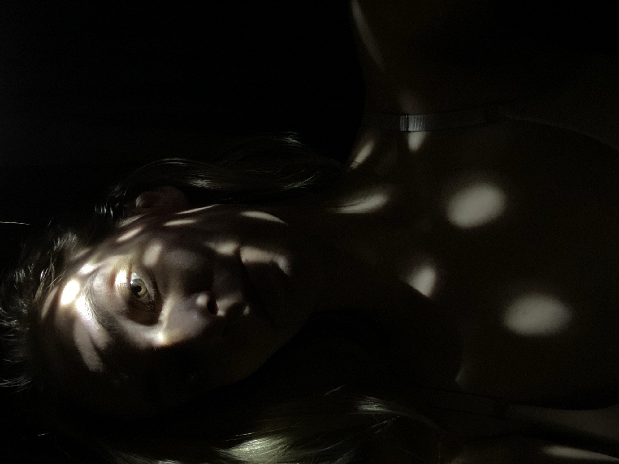 A portrait of a fair-skinned person with light hair, partially obscured by shadows. The person is lying down with their face tilted towards the viewer. Light patches, possibly from sunlight filtering through a tree or patterned screen, fall across their face and upper body. They are wearing what appears to be a silver chain around their neck. The background is predominantly dark. The location is unknown.