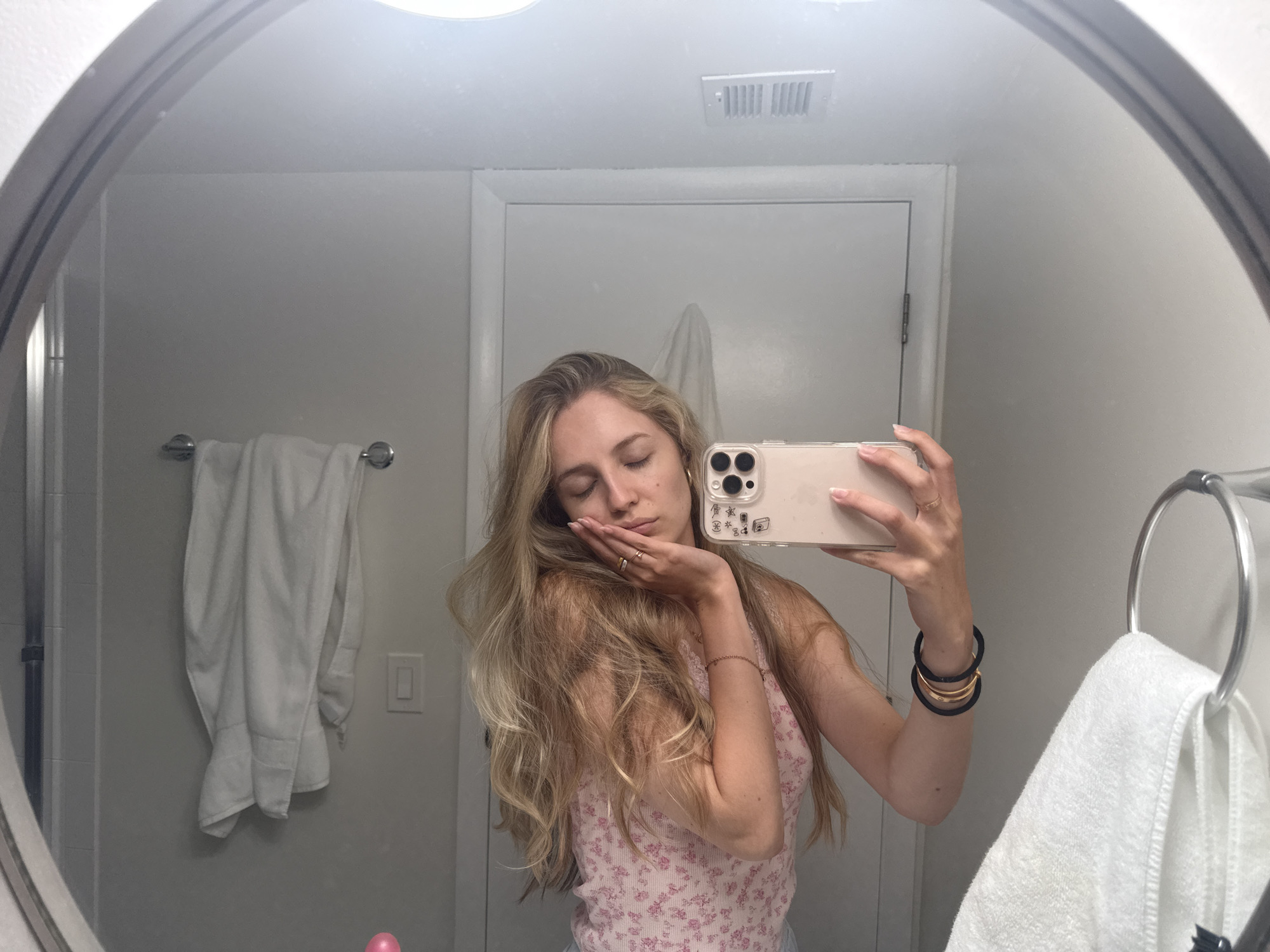 A young Caucasian woman with long blonde hair is taking a mirror selfie in a bathroom. She is wearing a pink floral tank top and has her eyes closed, with her head resting on her hand. She has on rings and bracelets on her wrists. In the background, there is a white towel hanging on a rack, a door, and a vent on the ceiling. The scene appears to be in a residential bathroom, likely in Miami, Florida.
