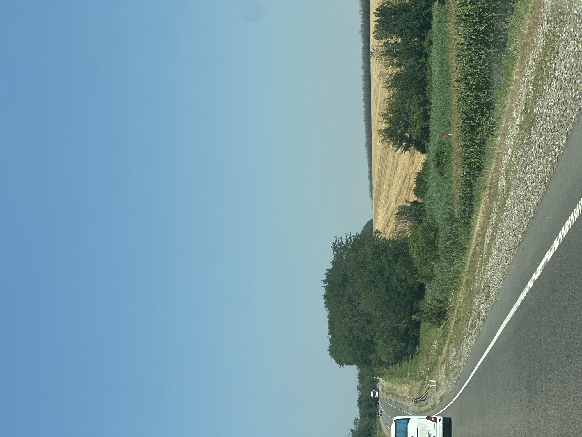 An image shows a highway with a white bus in the foreground on the left side. The bus is traveling on a road that curves to the left. To the right of the road, there is a field of dry straw and a field of corn. In the background, there are trees and a clear blue sky. The scene appears to be in a rural area.