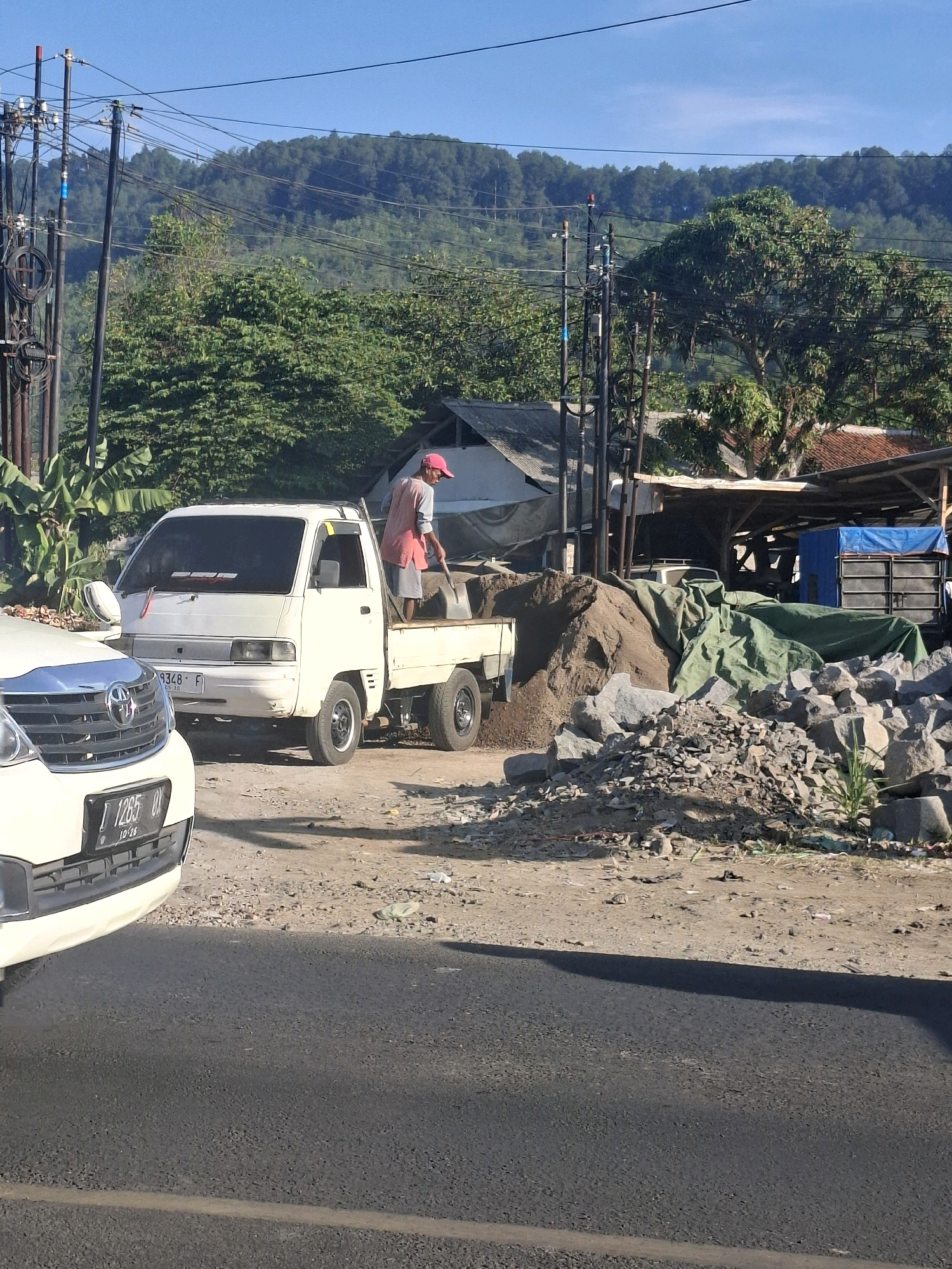 The image depicts an outdoor scene in Lembursawah, Indonesia, with a clear blue sky and a verdant, tree-covered hillside in the background. In the foreground, a white pickup truck is parked on a dirt road. A person wearing a pink cap and a reddish-pink top is shoveling sand from the truck bed into a pile. To the left of the pickup truck, the front portion of a white car is visible. The ground is covered with dirt, gravel, and piles of sand and rocks. Various construction materials and tarps are scattered around the area. Power lines and utility poles are prominent in the middle ground, suggesting a semi-urban or rural setting.