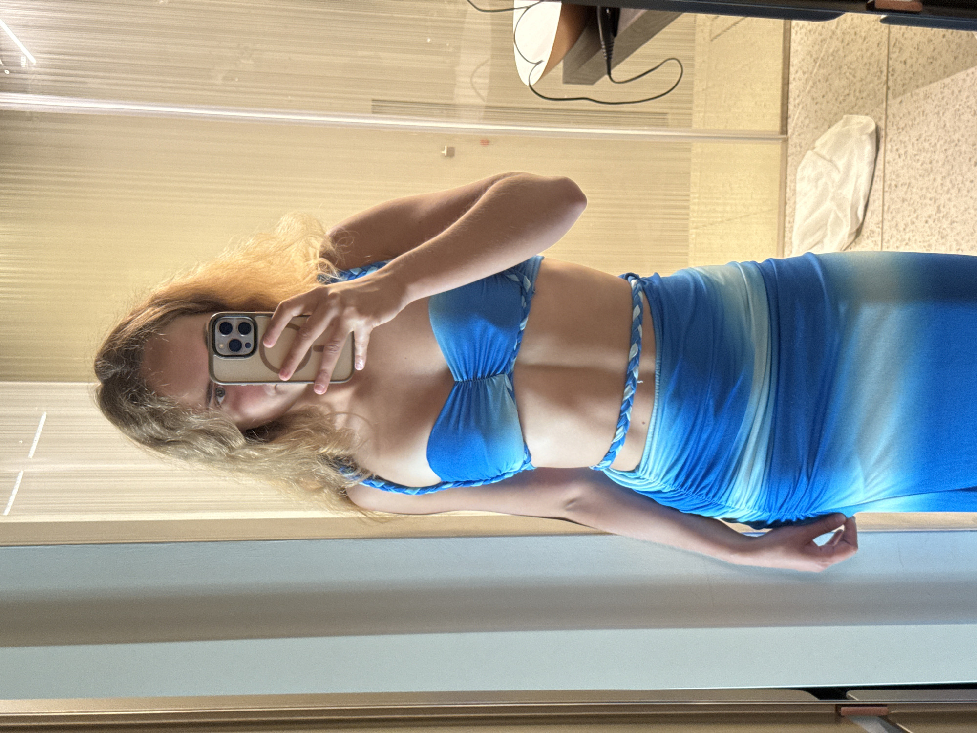 The image shows a young woman with blonde hair taking a mirror selfie indoors. She is wearing a blue ombre two-piece outfit consisting of a ruched bandeau top with braided straps and a ruched maxi skirt with a braided waist detail. The background appears to be a room with a light-colored wall and a door with vertical fluting. A partial reflection of the room is visible in the mirror, including a table with a bowl and some cables. The floor is tiled with a speckled pattern. There is no indication of the city of Trieste, Italy in the image.