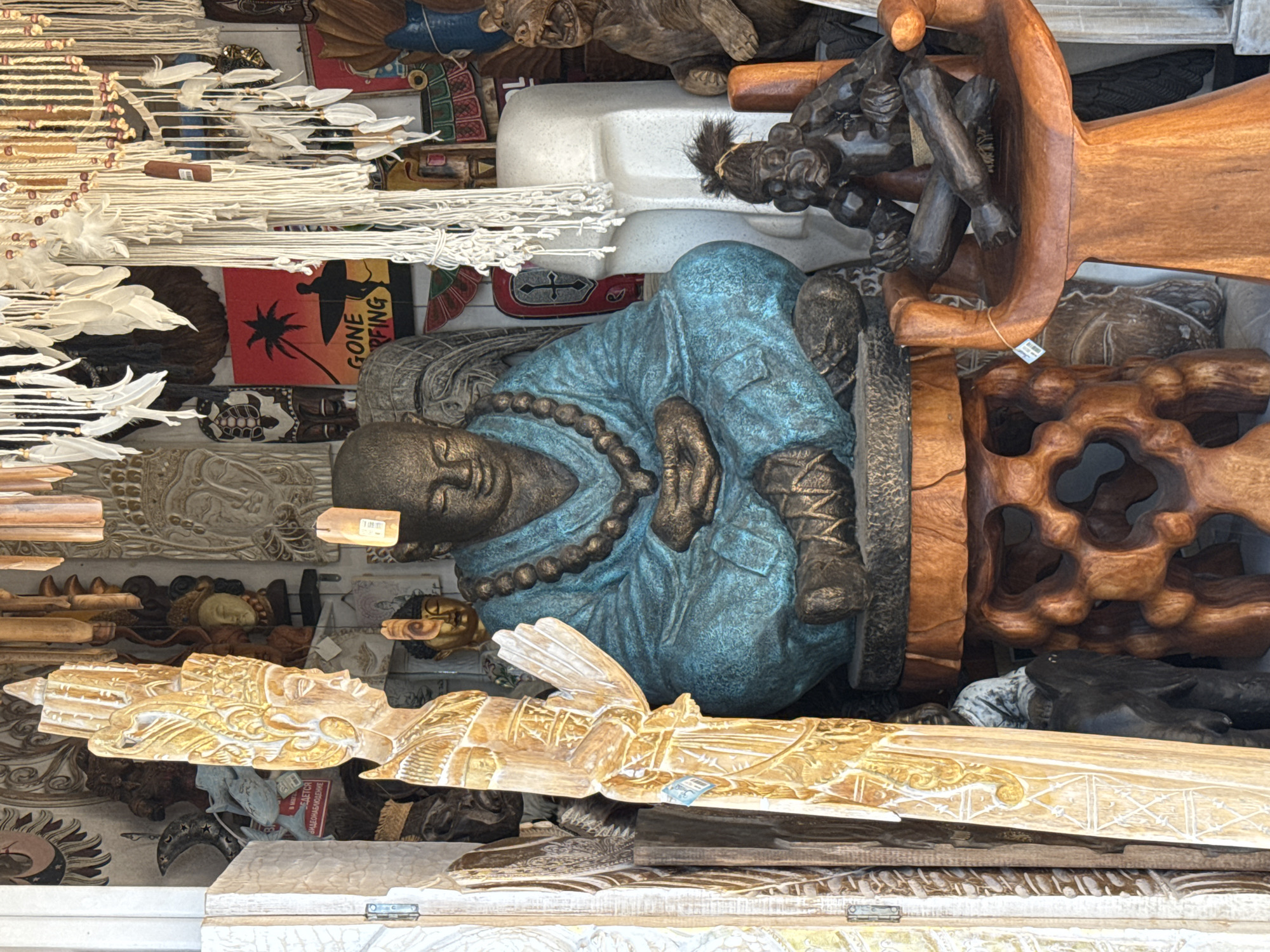 This media file displays a cluttered collection of various decorative objects, likely souvenirs or craft items, densely packed together in what appears to be a market stall or shop. The primary subject is a large, teal-colored statue of a meditating Buddha, positioned centrally and lying on its side. The Buddha statue is adorned with a beaded necklace and wears traditional robes.
Surrounding the Buddha are numerous other items, including carved wooden figures, some depicting human or animal forms, and intricately designed wooden panels. There are also strings of white beads and feathers, possibly part of dreamcatchers or similar decorations. Among the clutter, a small, red sign with a black silhouette of a surfer and a palm tree is visible, bearing the text "GONE SURFING". Another small, rectangular tag with what appears to be a barcode is attached to some of the items. The overall impression is one of an eclectic display of artisanal crafts. The lighting suggests an indoor setting, possibly illuminated by artificial light, as there are no clear indicators of time of day or weather conditions. The location context provided is Sochi, Russia, which might suggest the origin or style of some of the items.