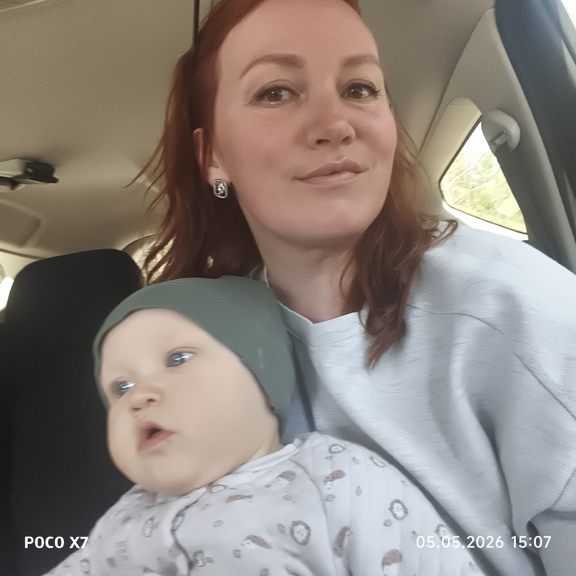 A woman and a baby are in the passenger seat of a vehicle. The woman has red hair, fair skin, and is wearing a light blue top. The baby is wearing a grey hat and a white onesie with a printed pattern of animals. The woman appears to be holding the baby. The photo was taken with a POCO X7 on May 5, 2026, at 15:07. The image's location is Chaikovsky, Russia.