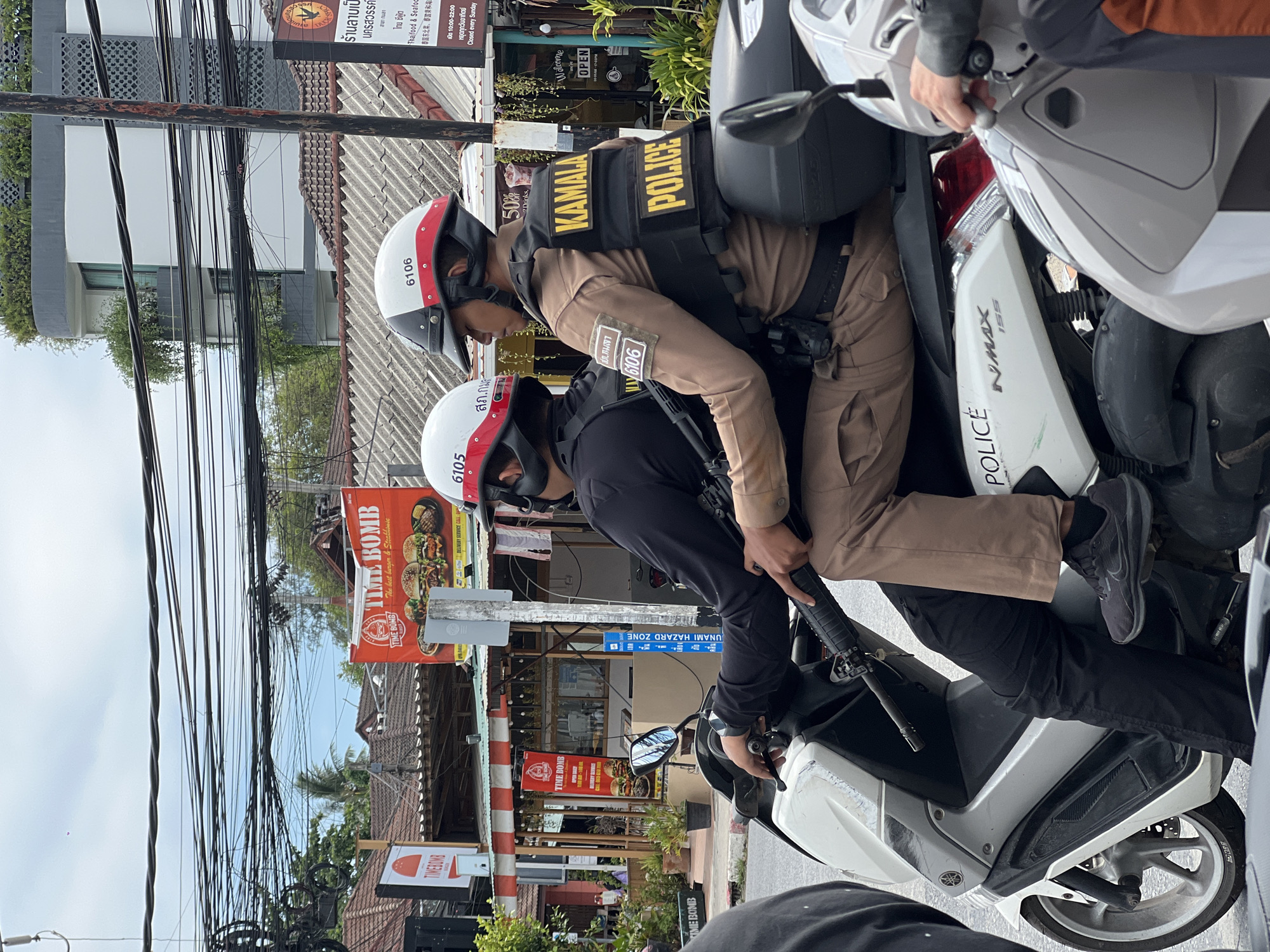 Two police officers are on motorcycles in Kamala, Thailand. Both officers are wearing helmets and body armor with "KAMALA POLICE" printed on the back. One officer wears a tan uniform, the other wears black. The rear motorcycle has the word "POLICE" printed on it, and "NMAX 155" is printed on the side. In the background, there is a sign for "TIME BOMB The best burgers & Steakhouse", and "UNAMI HAZARD ZONE". A restaurant sign says "ร้านลาบเป็ด". There are numerous overhead electrical wires.