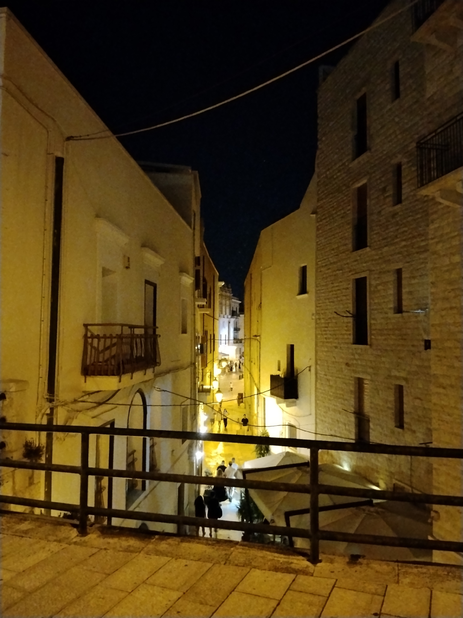 This is a nighttime photograph taken from an elevated perspective, looking down a narrow street in Bari, Italy. The street is lined with buildings made of light-colored stone and stucco, illuminated by artificial yellow lights. The sky above is dark blue. In the foreground, a dark metal railing runs horizontally across the image, with a tiled pathway in front of it.
The street itself is populated with a few people walking. Some are silhouetted against the light, making them difficult to discern. One group of people, appearing to be two women and a man, are walking away from the camera in the middle ground. Further down the street, more individuals are visible, suggesting a bustling evening atmosphere.
On the right side of the street, a multi-story building features windows and balconies. The building on the left is also multi-story, with a balcony visible. In the distance, the street curves and appears to open up into a wider area, with more buildings and lights. The overall impression is of a charming, historic European street scene at night.