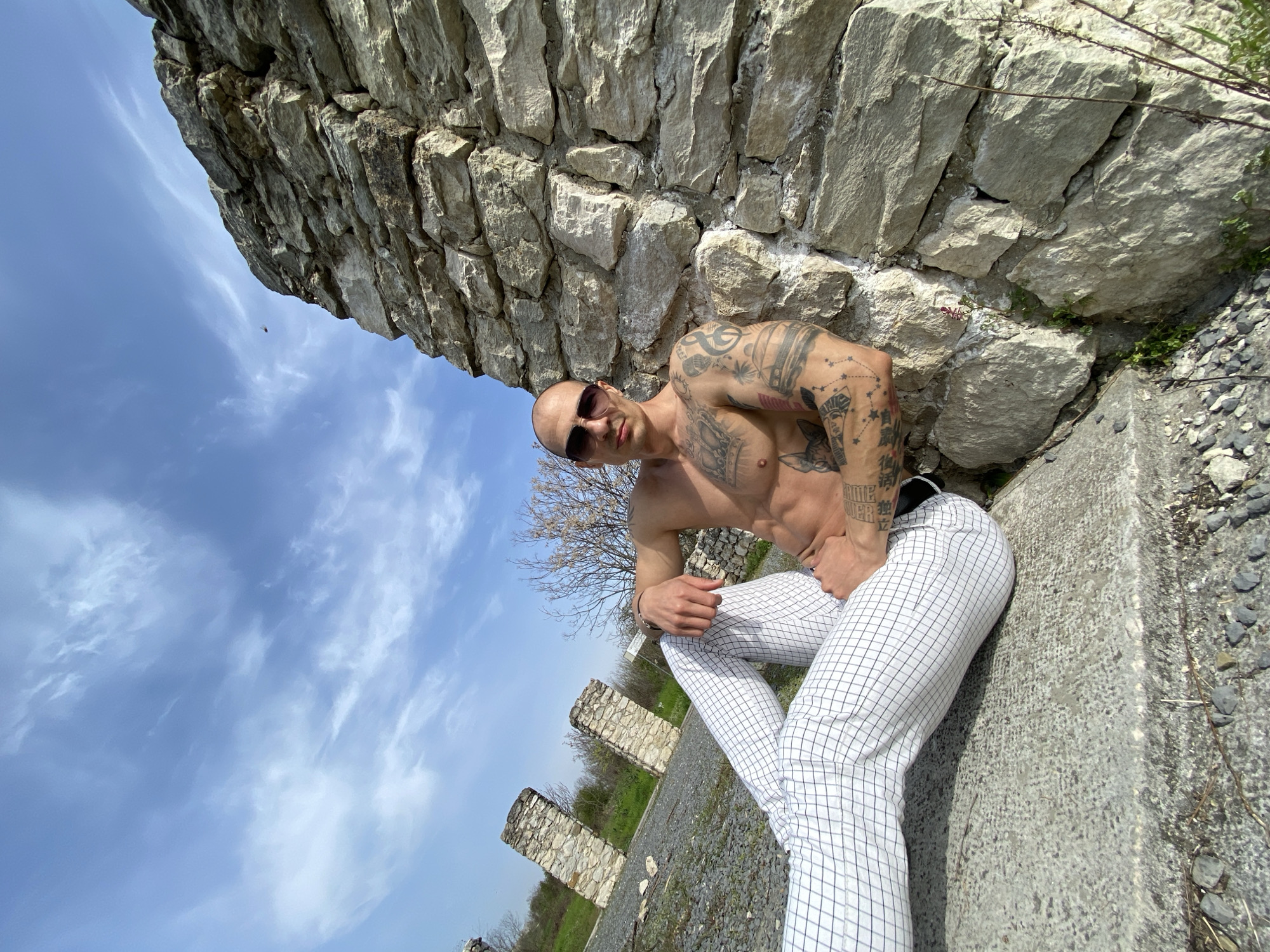 A shirtless man wearing white checkered trousers and sunglasses is leaning against a stone wall. He has numerous tattoos on his arms and torso. He is sitting on a concrete structure. In the background, there is a blue sky with clouds and some stone pillars. Based on the style of the stone walls and pillars, this scene appears to be located within or near the ruins of the Kaylaka Park in Pleven, Bulgaria.
