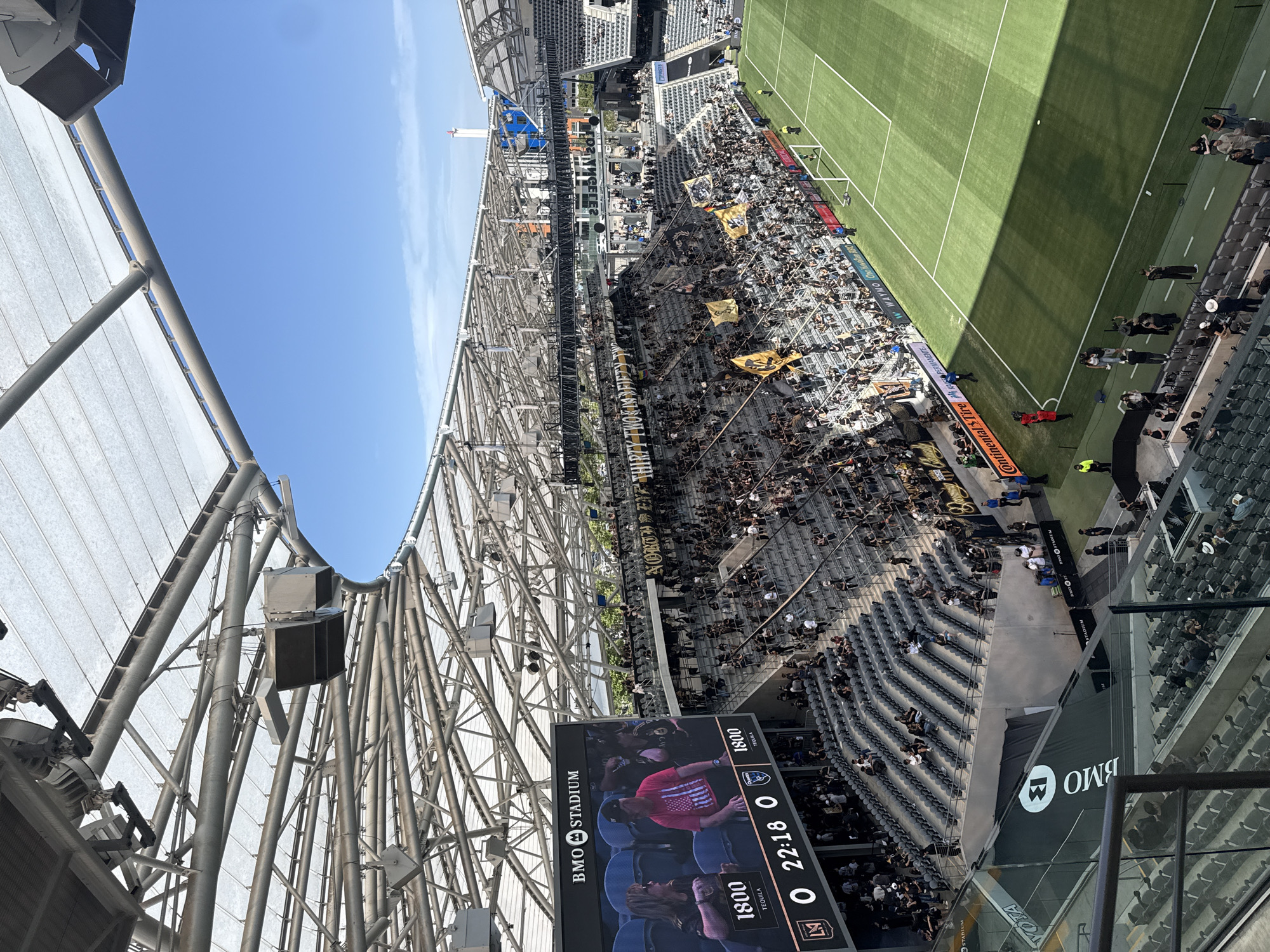 Here is a concise description of the image:
The image shows an elevated view of BMO Stadium, a soccer-specific stadium in Exposition Park, Los Angeles, California. The stadium is filled with spectators; many are wearing black and gold colors, and some are holding yellow flags. A soccer match is taking place on the green field. A large display screen in the foreground shows the score and game time. The stadium is covered by a large structural roof. The weather appears to be sunny, with a clear blue sky visible.