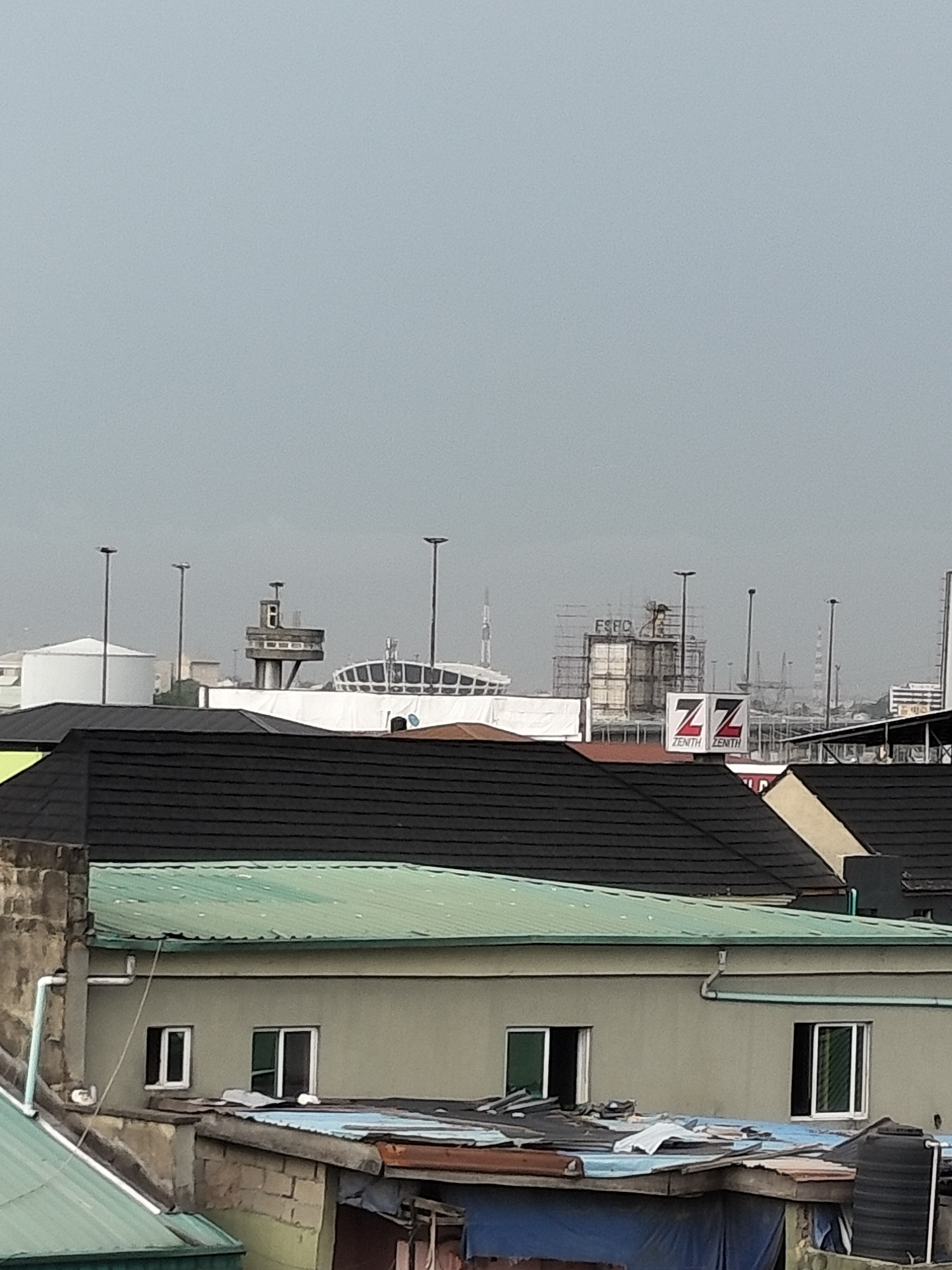 The image shows a view of Lagos, Nigeria on an overcast day. In the foreground, there are rooftops of residential buildings with varying materials - black tiles, green metal sheets, and rusting corrugated metal. The houses are a mixture of cement and other materials. In the background, industrial facilities, including storage tanks, control towers, and possibly part of a stadium, are visible. There is a large sign with the letters "ZZ" and the word "ZENITH" twice on it. The sky is uniformly gray.