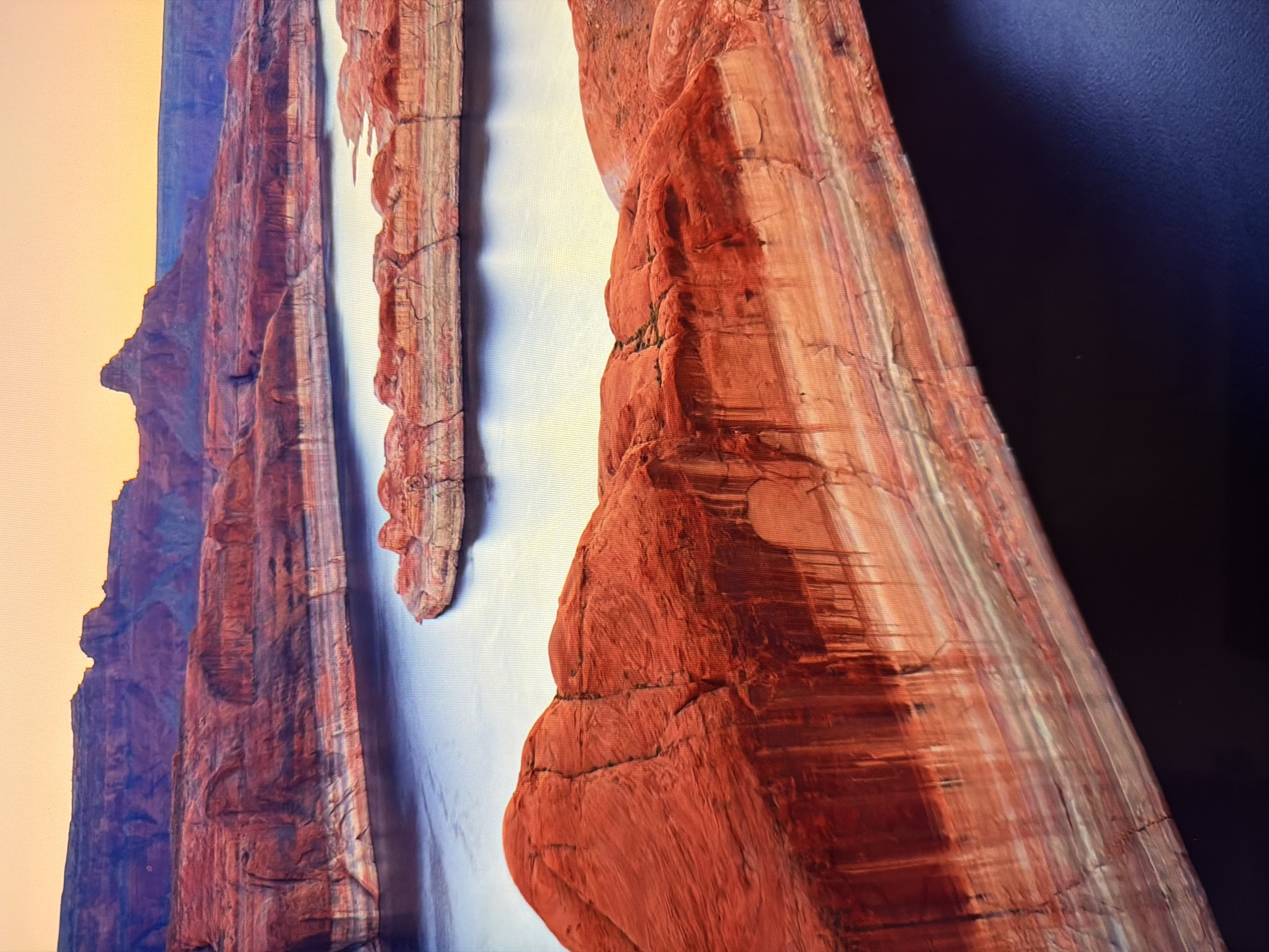This image depicts a vast, arid landscape with layered red rock formations and a body of dark water. The rock strata exhibit a variety of colors, including deep reds, oranges, and lighter bands of beige and white. The water appears still and reflects the warm tones of the sky and surrounding rock. In the distance, the landscape continues with more red rock formations under a pale orange sky. There are no signs of human presence, urban environments, or specific landmarks that would place this scene in Kyiv, Ukraine. The environment appears natural and undeveloped.