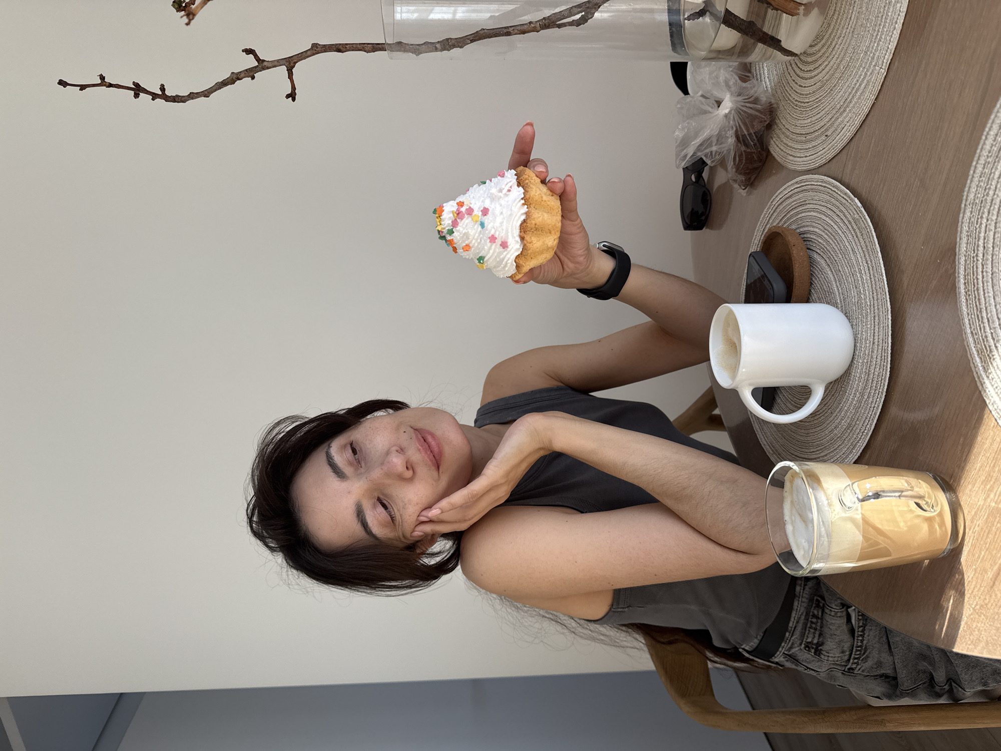This image, rotated 90 degrees clockwise, captures an indoor scene in Perm, Russia, featuring a young woman enjoying a snack and drink at a wooden table. The woman, positioned on the left side of the frame, has dark hair and a fair complexion with some freckles. She is wearing a dark grey tank top and dark jeans, looking towards the viewer with a gentle smile and her right hand softly touching her face.
In her raised left hand, she holds a cupcake with white whipped frosting generously adorned with colorful, star-shaped sprinkles. On the wooden table to her right, two round, light-colored woven placemats are visible. On the lower placemat, there's a white ceramic mug filled with a coffee drink topped with foam. Beside the mug, a cork coaster and a small dark object are present. Further to the right and slightly below, a clear glass contains a layered coffee drink, also with foam.
The background consists of a plain, light-colored wall. Above the table on the upper right, a clear glass vase holds a bare, branched twig as a decorative element. A small transparent plastic bag containing dark items is also visible near the vase. The bright, even lighting suggests it is daytime. The woman appears relaxed and content. There is no visible text in the image.