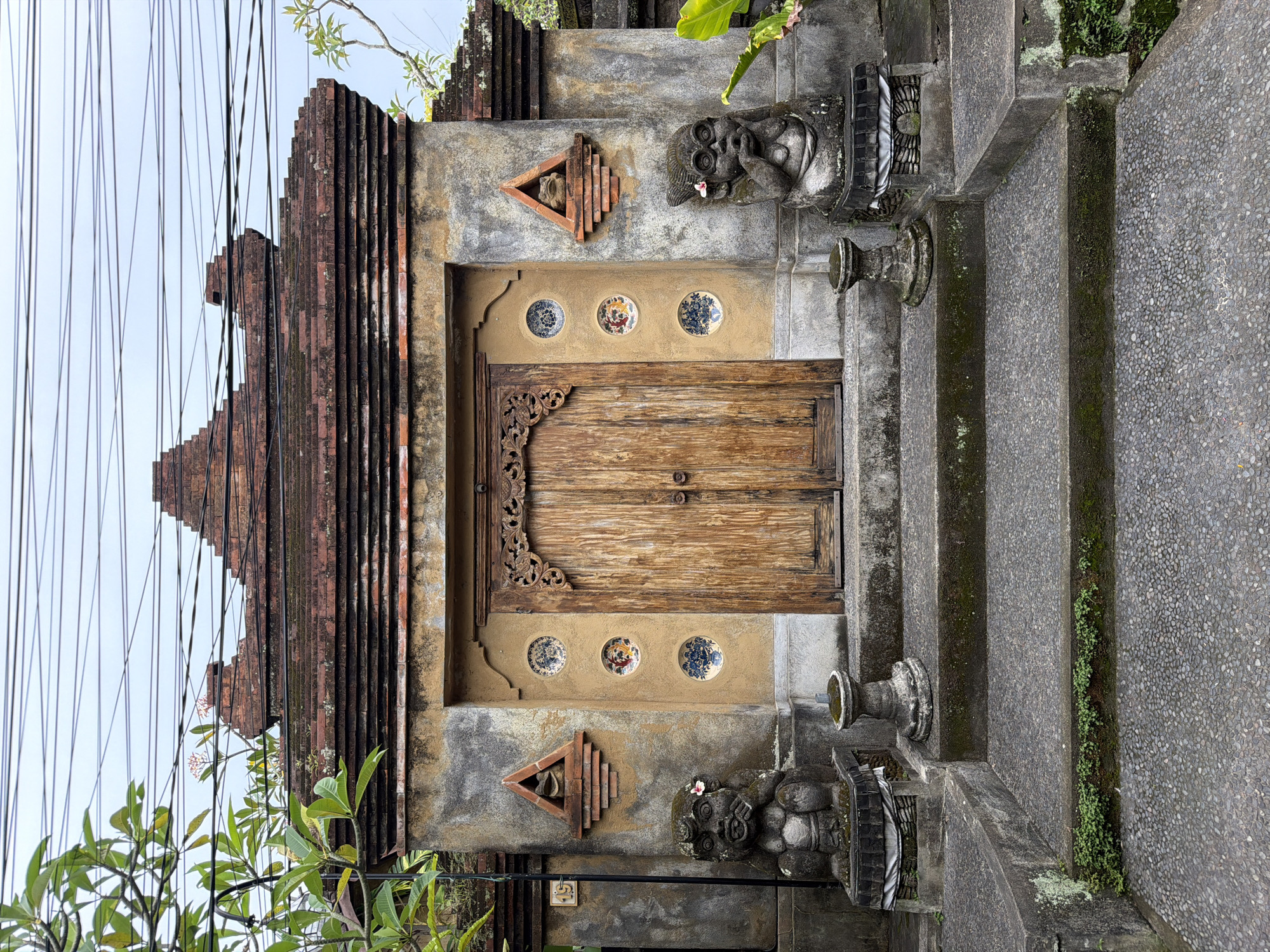 The image captures the elaborate, traditional entrance to a building, likely a temple or a traditional home, in Ubud District, Indonesia. The composition is centered on a grand, ornate wooden double door, featuring intricate carvings, particularly along its upper frame and handles.
Above the wooden door, set within a lighter, textured wall section, are three circular decorative medallions or ceramic plates, each displaying unique floral or abstract patterns. Below these, the wall is a darker, weathered concrete. Flanking the entrance steps, on elevated platforms, are two intricately carved, moss-covered stone guardian statues. These figures possess distinct, stylized facial features, with one appearing to hold its hand to its face, and both are adorned with small, delicate pink flowers.
The entrance is approached by a series of grey concrete steps with a pebbled texture. Moss and small green plants grow in the crevices and along the edges of these steps, indicating a humid environment. Above the entrance, a distinctive multi-tiered, stepped roof structure made of reddish-brown bricks or tiles ascends, characteristic of traditional Balinese architecture.
To the left, numerous black electrical wires run vertically across the upper part of the frame, partially intersecting the roofline. Green foliage, including leaves and some small white and pink blossoms, is visible on both sides of the entrance and peeking over the top, providing natural accents. The sky overhead is a bright, overcast white, suggesting daytime conditions. There are no people visible, nor any discernible text.