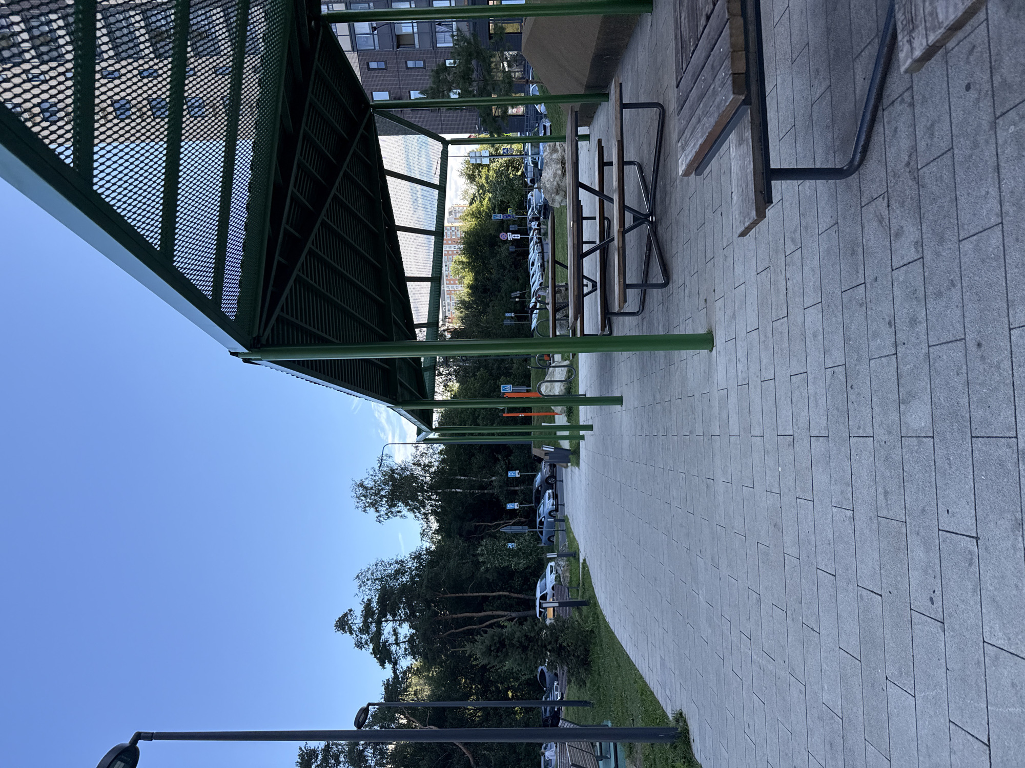 This is a photograph of an outdoor seating area with picnic tables under a green metal-framed shelter. The shelter has a perforated roof and is supported by green pillars. In the foreground, there is a paved walkway made of gray rectangular tiles. Several wooden picnic tables with benches are visible. To the left of the walkway, there is a grassy area with a bench and a lamppost. In the background, there is a parking lot with several cars parked, a dense line of trees, and modern apartment buildings. The sky is clear and blue. The scene appears to be in the city of Tomilino, Russia, possibly in a residential or park area.