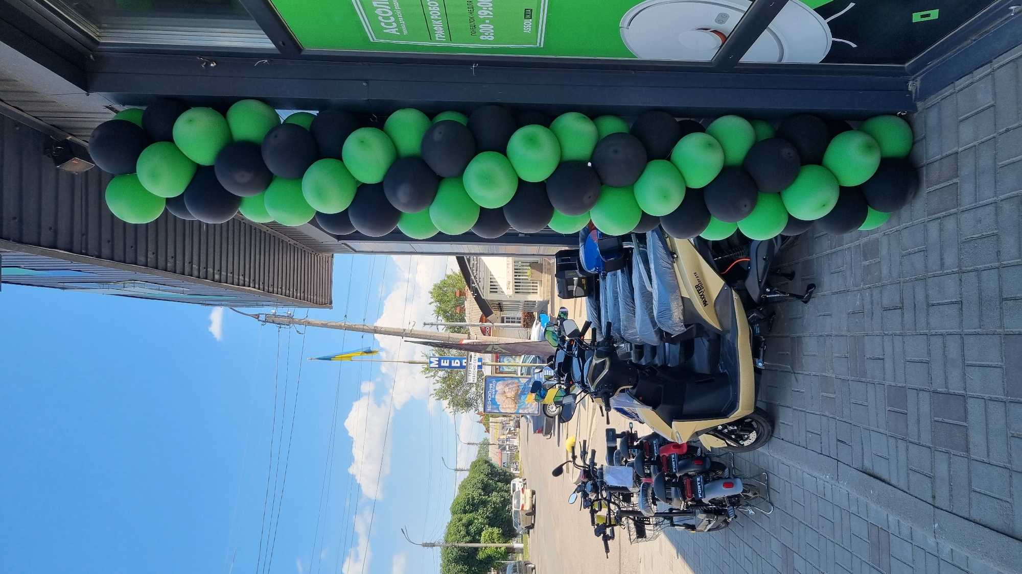 This media file captures an outdoor scene in Smila, Ukraine, likely on a bright, sunny day with a clear blue sky and scattered clouds. The foreground is dominated by a row of parked electric scooters, many of which are covered in plastic. These scooters are arranged on a gray paved surface.
Above the scooters, a festive decoration of black and green balloons stretches across the building's facade, suggesting a celebration or promotional event. The building itself appears to be a commercial establishment, with windows and signage visible. One sign, partially visible and bearing Cyrillic text, reads "АССОЛ" and provides information about business hours: "ПОНЕДІЛОК-НЕДІЛЯ 8:00-19:00". This translates to "MONDAY-SUNDAY 8:00-19:00", indicating the establishment's operating schedule. Another visible text, "MES", is part of a larger sign above the scooters. A Ukrainian flag, yellow and blue, is also visible flying from a pole in the background.
In the middle ground, a street with some traffic, including cars and more scooters, is visible. Trees and other buildings line the street, providing context of a town or city setting. The overall impression is one of an active commercial area decorated for an event, possibly a grand opening or sale, with a focus on electric scooters. There are no people clearly visible in the image.