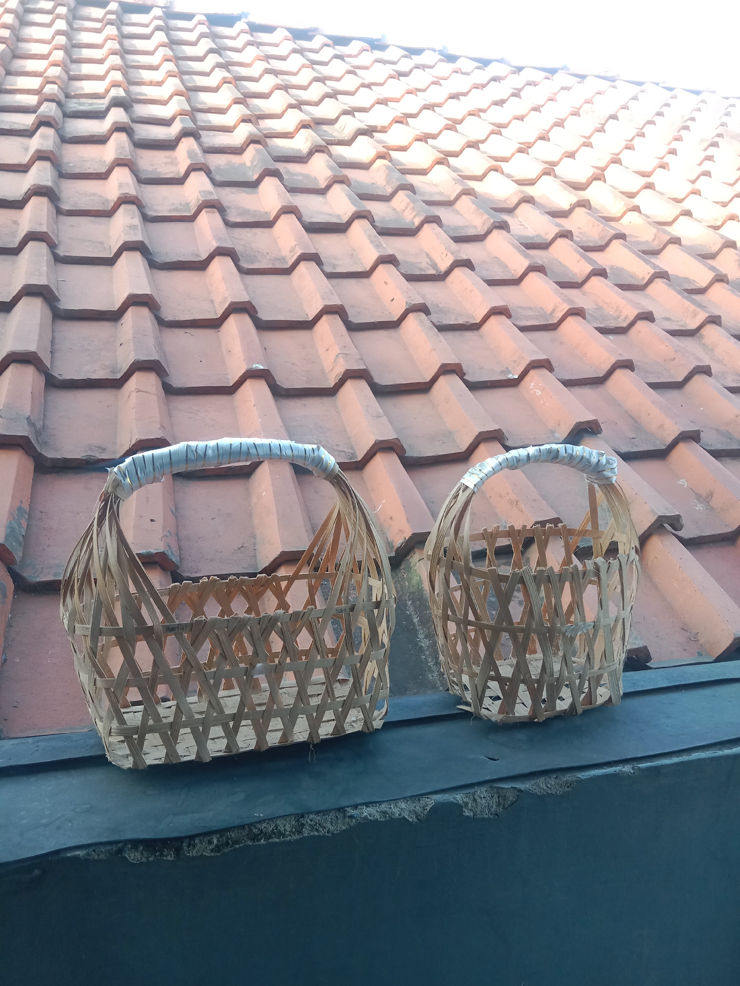 Two woven bamboo baskets with wrapped handles sit on a dark ledge in front of a tiled roof. The baskets are woven with a crisscross pattern. The roof tiles are reddish-brown and arranged in overlapping rows. The ledge appears to be made of concrete. The background is a bright, overcast sky. The image is taken from a slightly elevated perspective. The location is the city of Kutosari, Indonesia.
