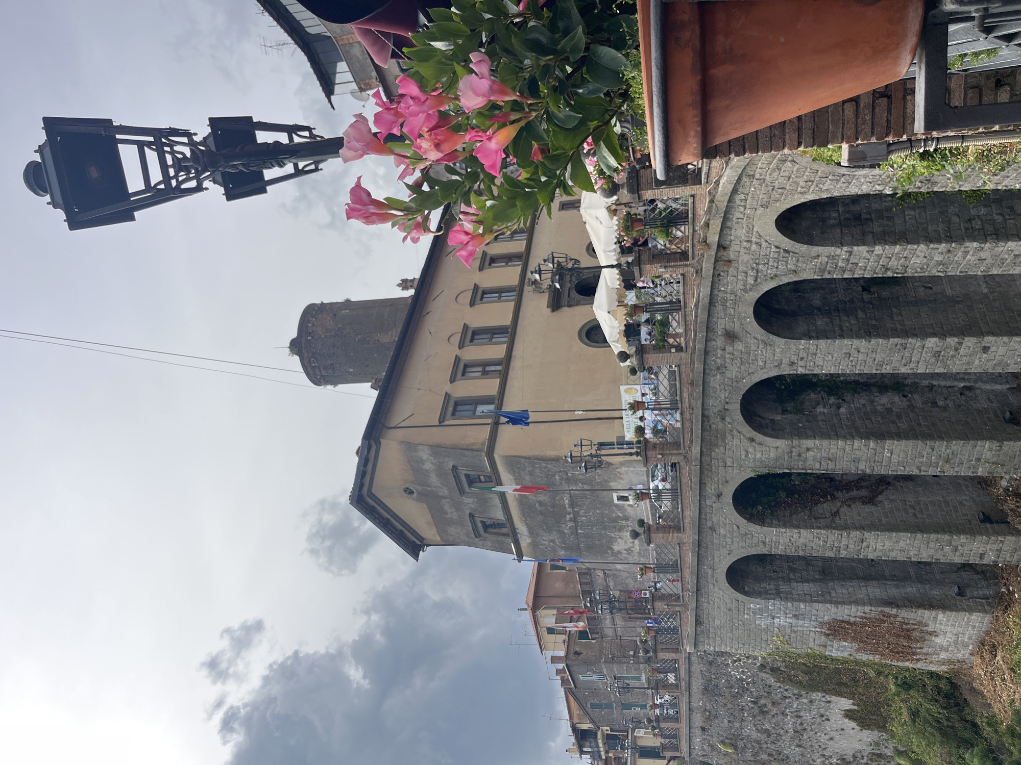 An outdoor, low-angle, medium shot captures a historic stone bridge with three arched openings, spanning a chasm. The bridge is topped with a railing and a walkway lined with potted plants and lampposts. Beyond the walkway, a light-colored, stucco building with a tower and multiple windows stands. Flags, including an Italian flag, are visible on poles in front of the building. The sky above is cloudy and overcast. In the foreground, on the right side of the image, a large terracotta pot filled with pink flowers and green leaves is visible. Buildings and a lamppost are also seen. The scene appears to be located in the town of Nemi, Italy, on a cloudy day.