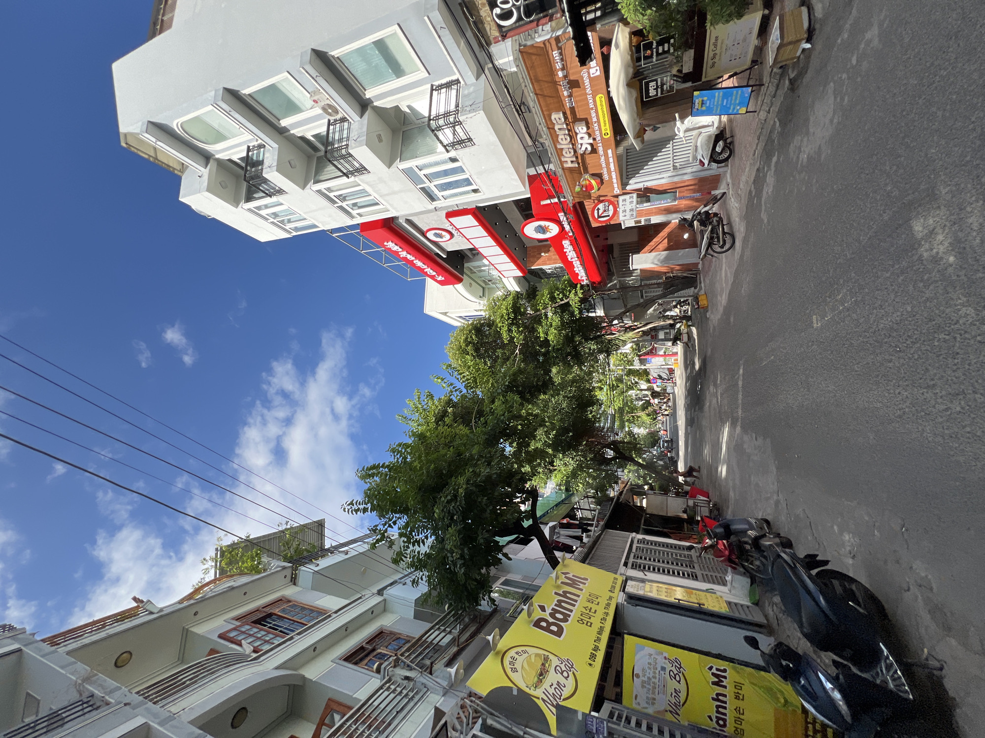 The image depicts a street scene in Nha Trang, Vietnam, on a clear day with a bright blue sky and scattered white clouds. The street is lined with multi-story buildings, some of which are painted in light colors like white and grey, while others feature vibrant red accents. Several businesses are visible, including a "Banh Mi" shop with a prominent yellow sign, a "Helena Spa" with brown signage, and a "Sip Sip Coffee" establishment with an open umbrella. Various types of motorcycles and scooters are parked along the sidewalks and street. Lush green trees are interspersed among the buildings, providing shade. In the distance, the street appears to continue, with more buildings and a few figures walking or riding vehicles. The overall impression is one of a typical urban street in a Vietnamese city, with a mix of commercial activity and residential buildings.