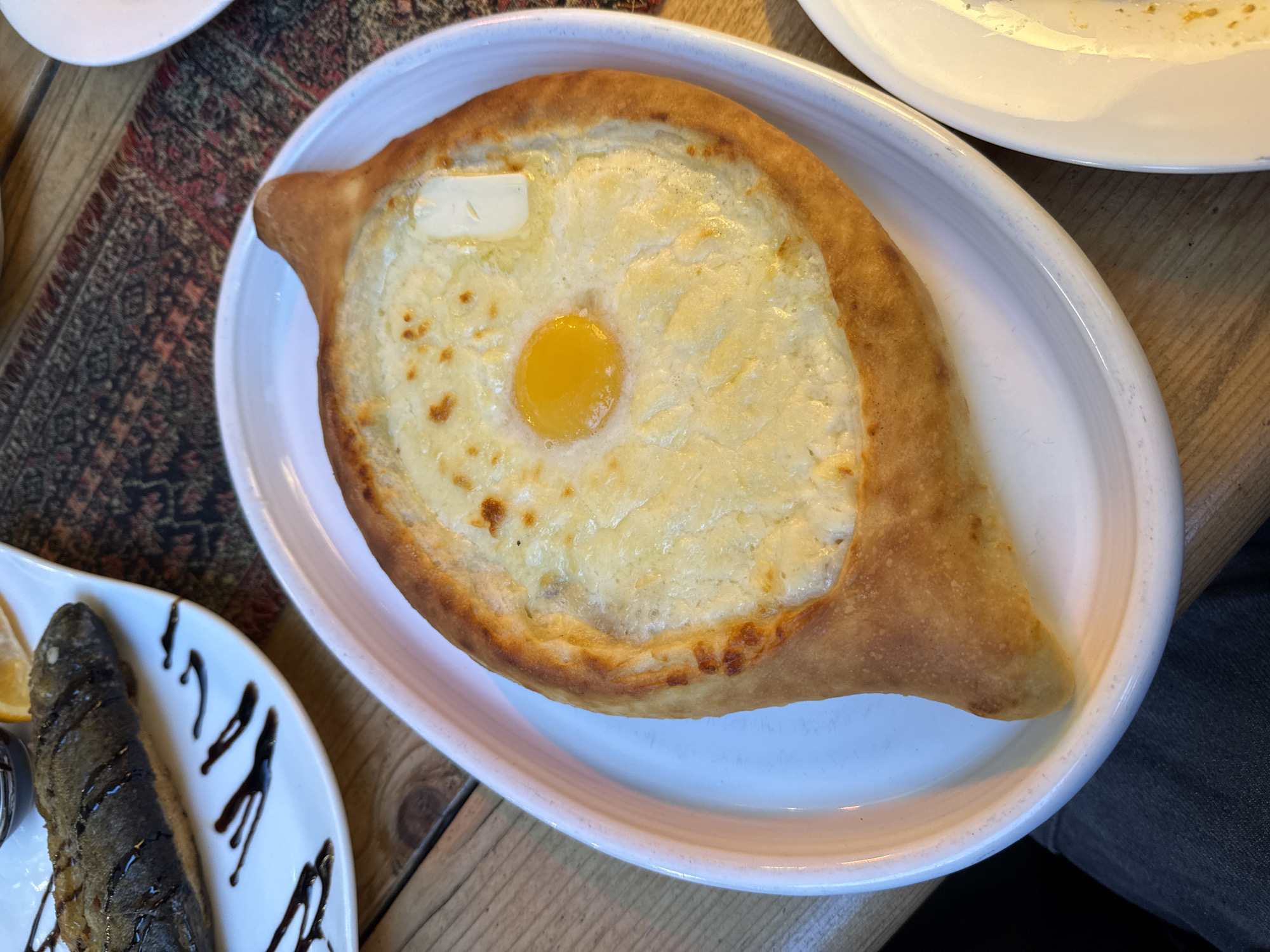 A flatbread, shaped like a boat, sits on a white oval plate, garnished with cheese, a raw egg yolk, and a pat of butter. The flatbread, known as Khachapuri, has browned crust. The cheese appears melted and bubbly. The raw egg yolk is positioned centrally on top of the cheese, and a single pat of butter is placed to its left. The dish sits on a wooden surface, possibly a table. A decorative carpet is partially visible to the left of the plate. Another plate, holding what appears to be grilled fish drizzled with a dark sauce, is to the lower left of the flatbread. Another plate is seen towards the top right. The scene is a restaurant setting, most likely in Tbilisi, Georgia, given the prominence of Khachapuri, a Georgian dish. No people are visible in the image, though the plate placement indicates they are likely seated at the table.