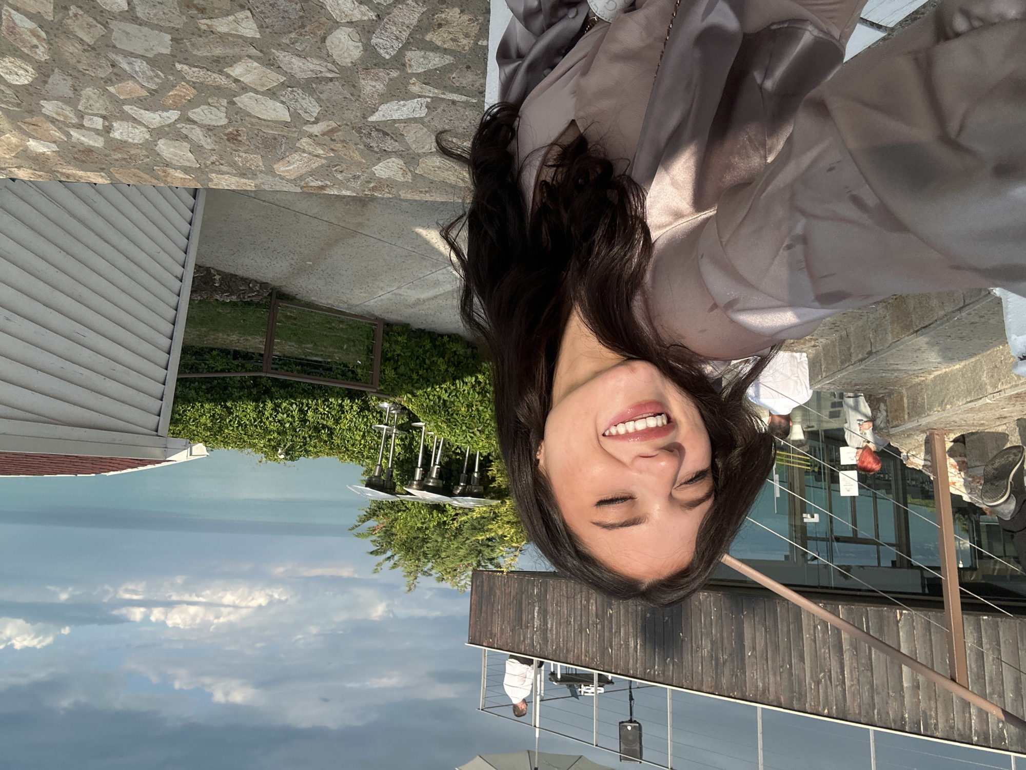 A woman with dark hair is taking a selfie outdoors. She's smiling with her eyes closed. She is wearing a taupe silky garment. The background shows a seascape with cloudy skies. There is a building with a wooden facade. There are other people in the background. There is also a house with white siding. The location is the city of Topola, Bulgaria.