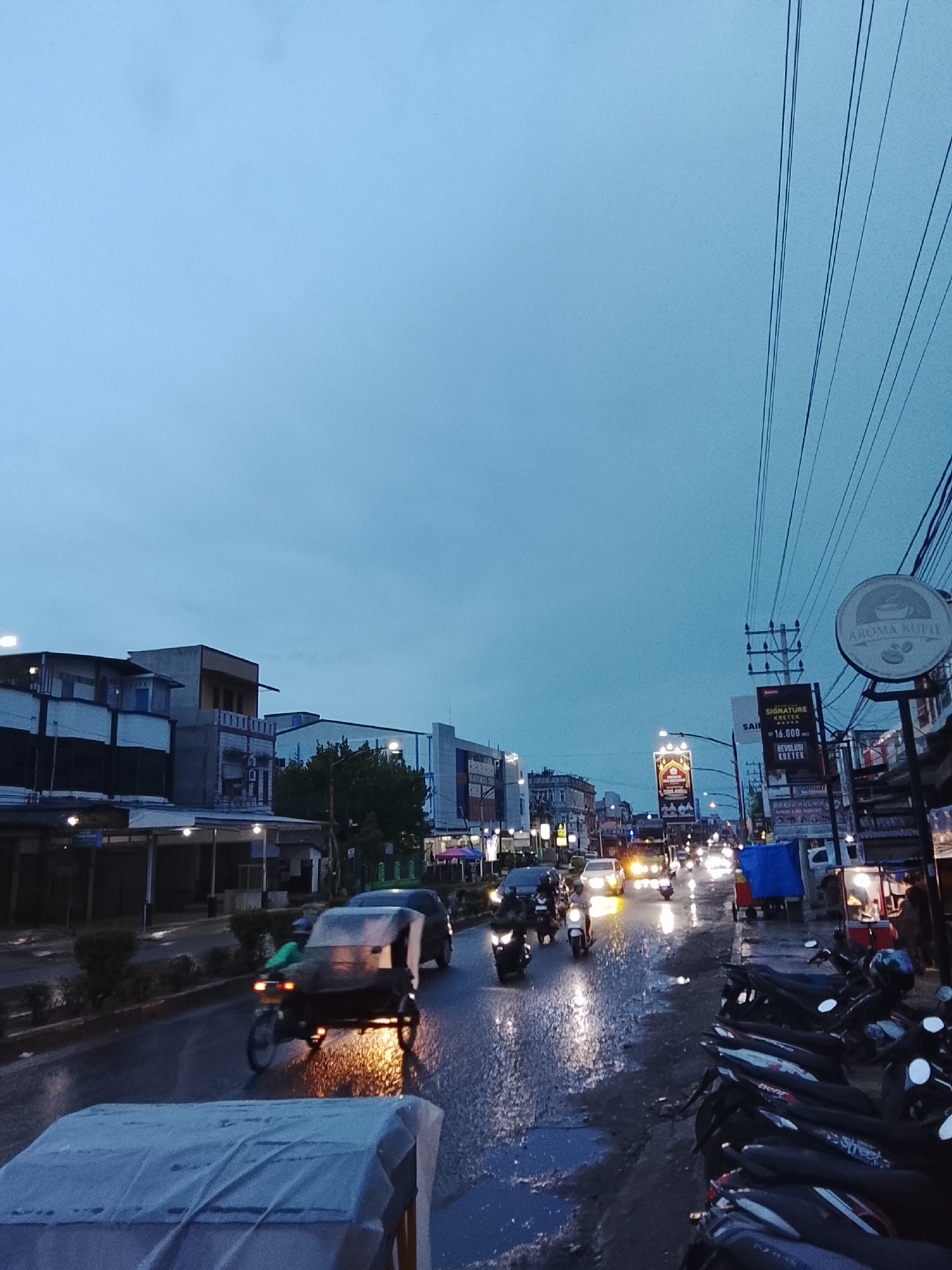 This image depicts an evening street scene in Lhokseumawe, Indonesia, characterized by the "blue hour" twilight and a wet road surface, suggesting recent rainfall. The sky is a deep blue, and artificial lights from streetlamps and vehicles are prominently reflected on the damp asphalt.
The street is busy with traffic. A three-wheeled vehicle, likely a local "becak motor" or "bentor," is central on the left side of the road, moving away from the viewer. A person in a green top is visible on this vehicle. Numerous motorcycles are present; some are parked along the right curb, while others are in motion on the road, their headlights illuminating the wet surface. Cars are also visible further down the street, their headlights shining brightly.
Buildings, varying in height and style, line both sides of the thoroughfare, some featuring illuminated commercial signs. Overhead, a dense network of power lines spans the sky on the right side. Several signs are legible: a prominent circular sign reads "AROMA KUPIH" (Aroma Coffee) with coffee bean graphics. Below it, a vertical banner advertises "SIGNATURE KRETEK" for "RP 16.000,-" (Indonesian Rupiah 16,000). Another sign below that mentions "BAKSO ARUMAN." The wet road and numerous puddles create a striking effect, enhancing the reflections of lights and giving the scene a vibrant, post-rain atmosphere at dusk.