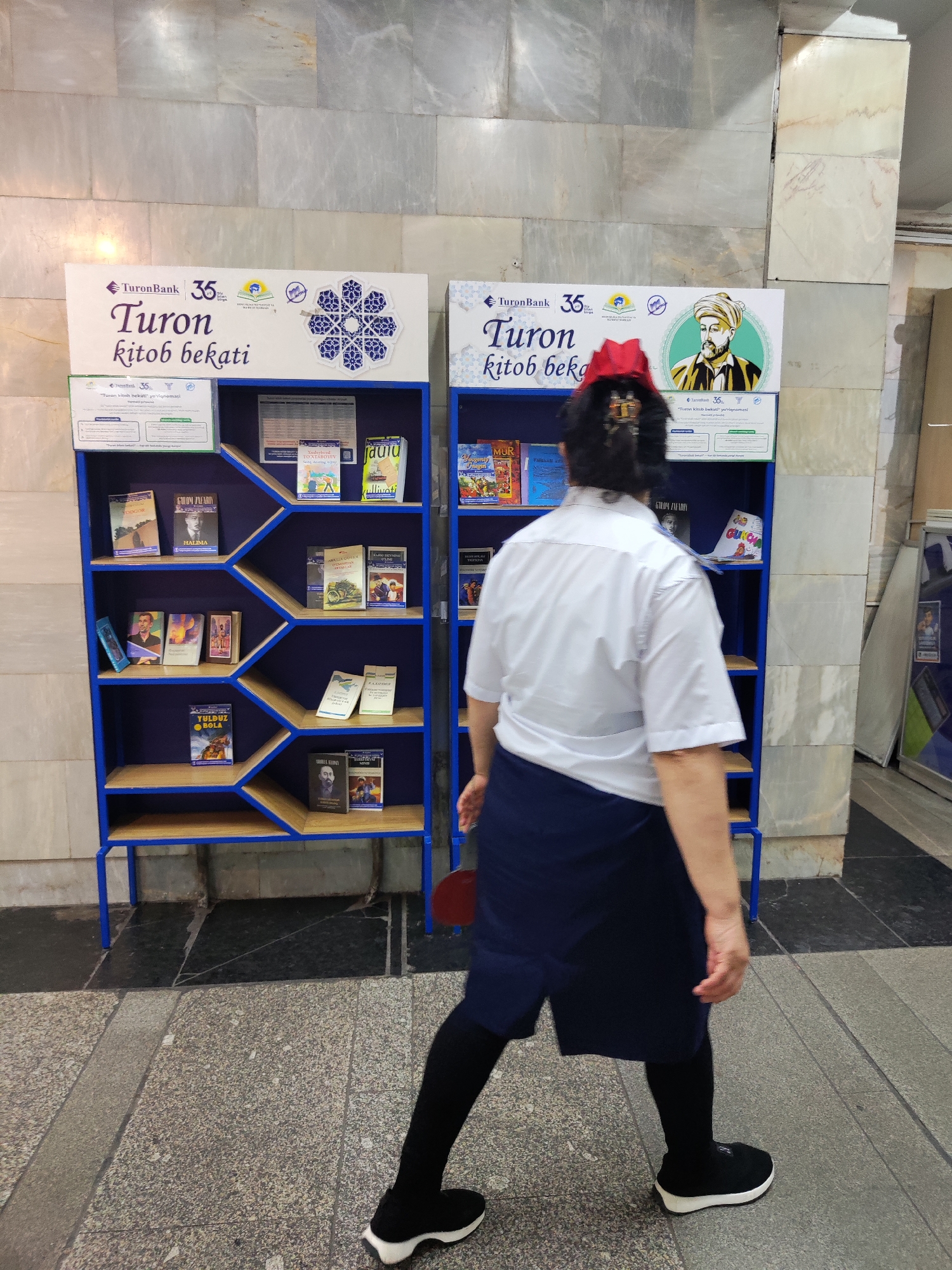 An individual wearing a white short-sleeved shirt, a navy blue skirt or apron over dark leggings, and black sneakers is walking past a blue bookshelf displaying books. The bookshelf is positioned against a tiled wall with grey and beige marble patterns. The bookshelf is labeled "Turon bank 35" and "Turon kitob bekati" in Uzbek. The individual has dark hair pulled back and is wearing a red hat. The scene appears to be an indoor public space in Tashkent, Uzbekistan, likely a bank or a library. The floor is tiled with a grey speckled pattern.