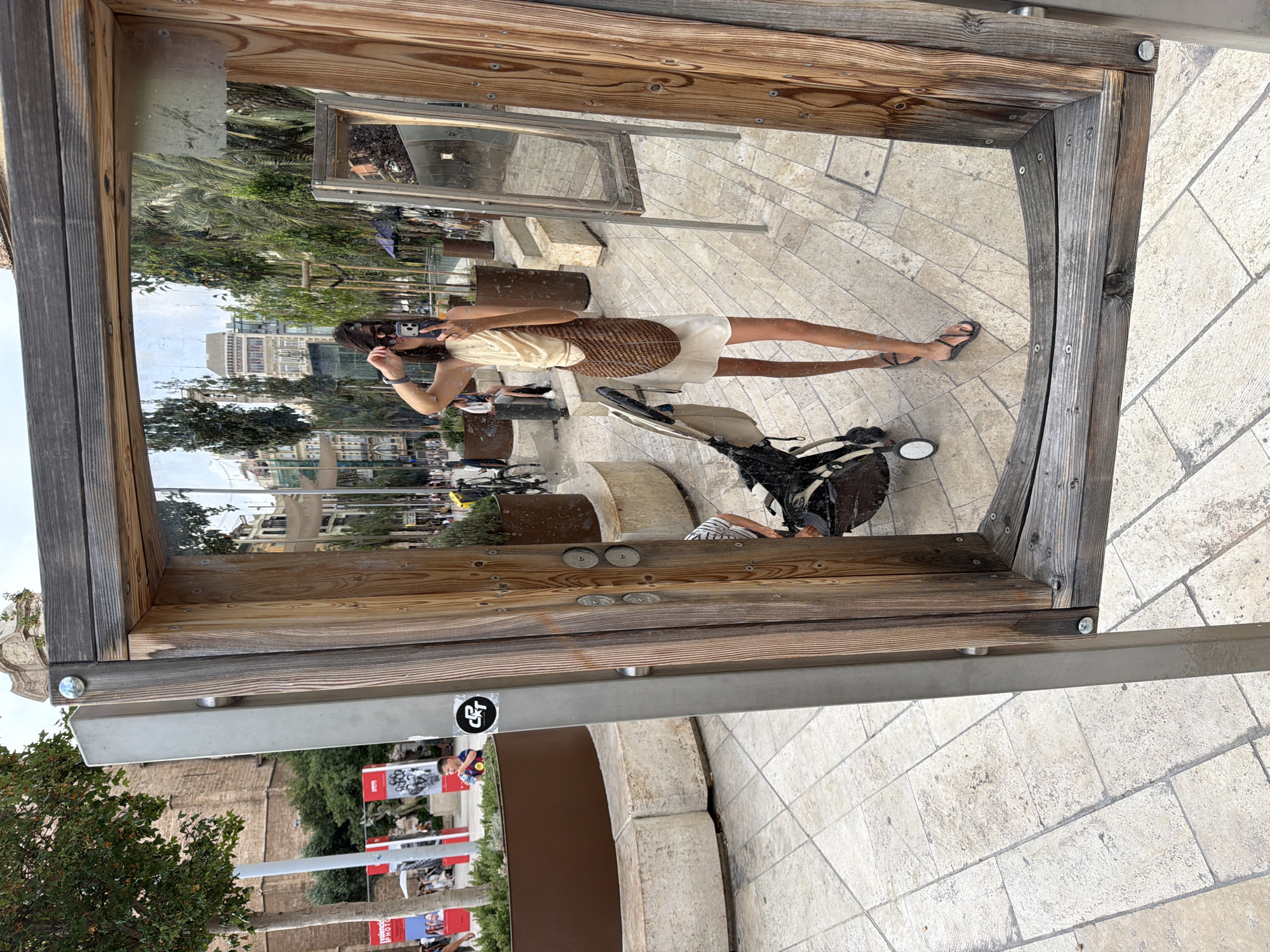 A young woman is taking a selfie in a large outdoor mirror with a wooden frame. She is wearing a light-colored, sleeveless halter top and a white skirt, and she has a woven bag slung over her shoulder. She is holding her phone in her right hand, with her left hand adjusting her sunglasses. Her feet are bare, and she is wearing flip-flops. Behind her, reflected in the mirror, is a city street scene with palm trees, buildings, and other people. In the foreground, to the left of the mirror, is a stroller. To the left of the mirror, outside the reflection, is a stone planter with greenery and a large tree. The ground is paved with light-colored tiles. The overall setting appears to be a public space in a city.