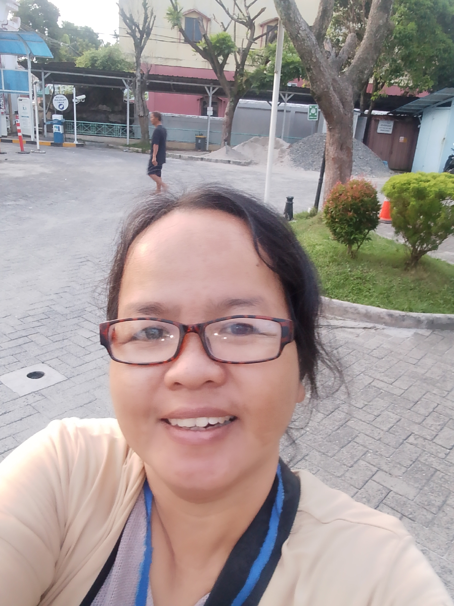 A medium shot selfie taken outdoors shows a woman wearing glasses and a light-colored jacket. She is smiling and looking at the camera. In the background, a man is walking away from the camera, and there are buildings, trees, and a gas station visible. The ground is paved with cobblestones. The image appears to have been taken in the city of Pematang Siantar, Indonesia, a location known for its mix of urban and natural landscapes. The woman's attire and the presence of a gas station suggest a public setting, possibly during daytime.