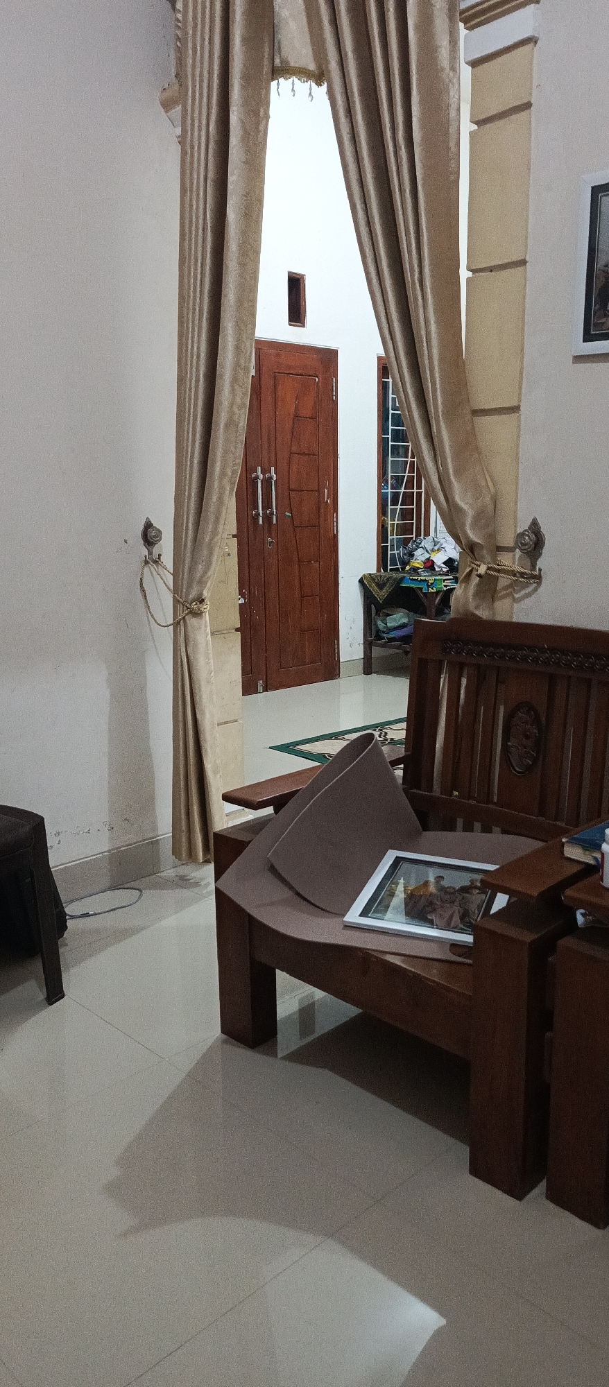 The image depicts an indoor scene, likely a residential interior, in Jatisari, Indonesia. The foreground features a wooden chair with a light brown cushion and a framed picture placed on it. To the left of the chair, a part of a dark brown object, possibly another piece of furniture, is visible. The floor is tiled in a light, neutral color.
In the mid-ground, a wooden double door is centrally located, with a small, dark rectangular window above it. To the right of the door, a window with a metal grille is visible, behind which some clutter is apparent on a small table. The room is partially divided by golden-brown curtains, which are tied back on the left side of the door. On the wall to the left of the door, a metallic hook with a rope tied to it is attached. The overall lighting suggests it is either evening or early morning. The architecture includes architectural columns and moldings.