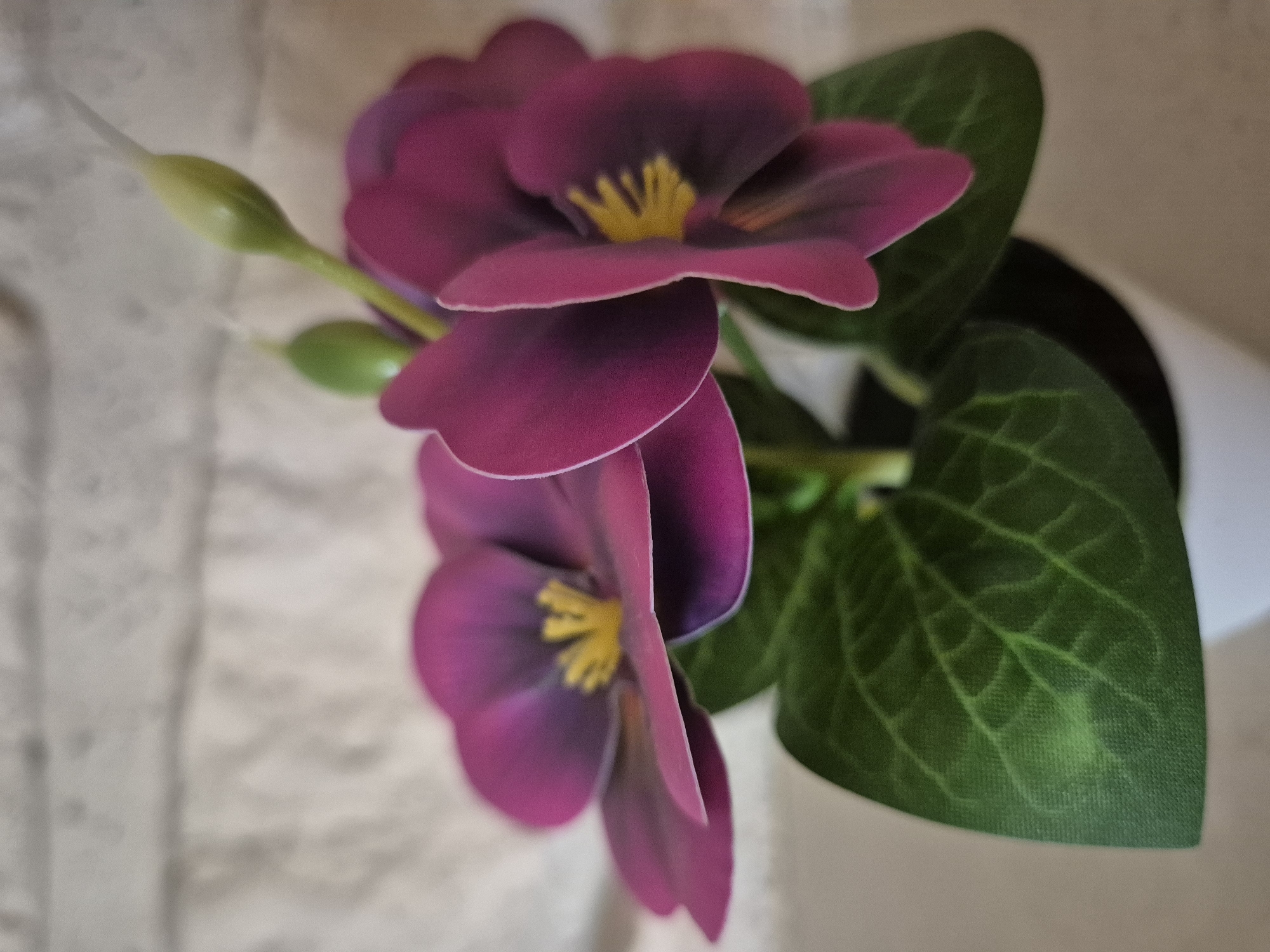 A close-up, shallow depth of field image shows a potted artificial plant with two vibrant purple pansy-like flowers and green leaves, set against a blurred light-colored brick background. One flower is in sharp focus, displaying its yellow center and layered petals. Another flower is slightly out of focus to the left. Two green buds are visible above the flowers. The plant is in a white pot, which is partially visible at the bottom of the frame. The overall impression is of a decorative item, likely placed indoors. The location is unknown.