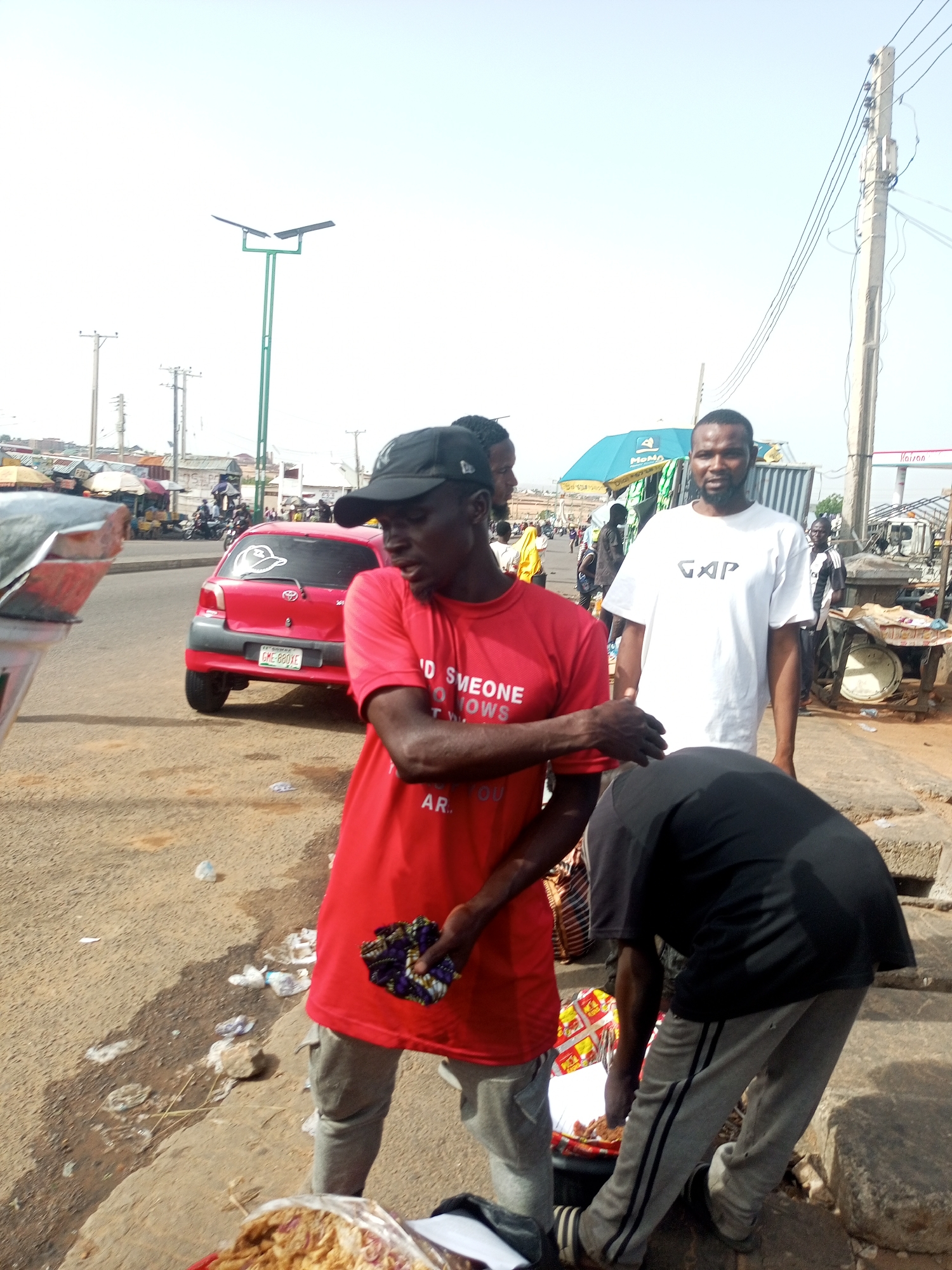 The image depicts a street scene in Gombe, Nigeria. Three men are in the foreground. One man wears a red t-shirt and a black cap and holds a colorful cloth. Another man stands behind him, wearing a white t-shirt. The third man bends over a container. A red car is visible in the background. The street is littered, and other people, vehicles, and vendors are present in the distance. Streetlights and utility poles are visible overhead.