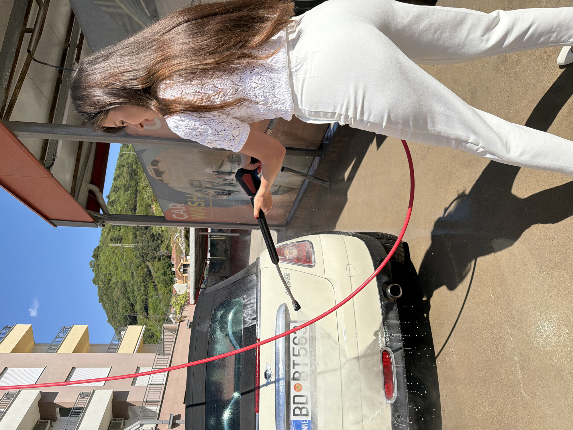 A woman washes a light-colored Mini Cooper car with a pressure washer in a car wash in Budva, Montenegro. She is wearing a white lace top and white pants. The license plate on the car is visible. Buildings and green trees are visible in the background. A red hose snakes from the pressure washer to the car. The scene is sunlit.
