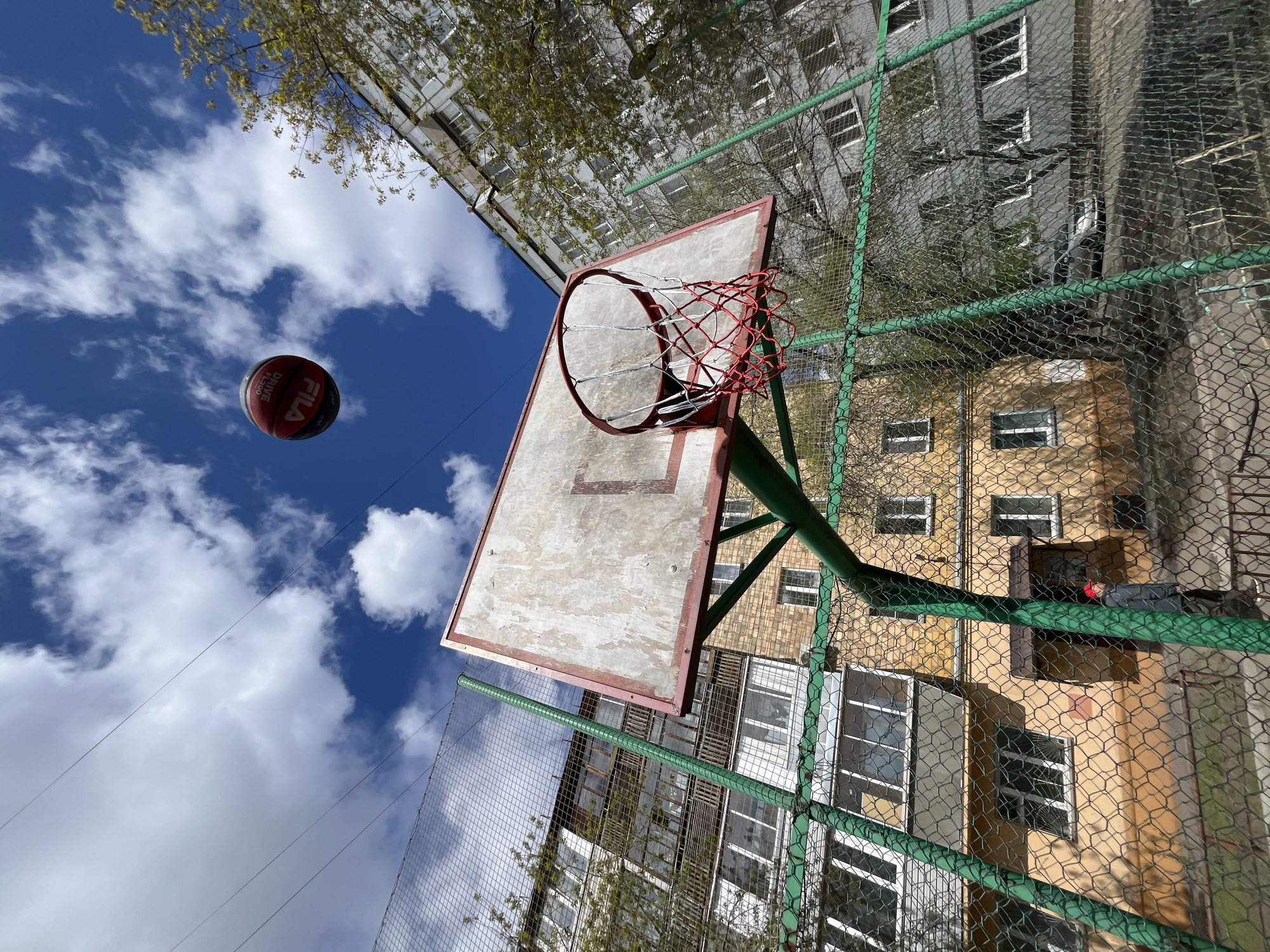 The image shows a basketball court located in an urban environment, likely in Krasnoyarsk, Russia. A basketball is suspended in the air against a blue sky with scattered clouds. The basketball hoop is visible with a worn backboard and a red net. A green metal fence surrounds the court. In the background, there are residential buildings with multiple windows, some showing signs of age and wear. A person can be seen laying down behind the fence.