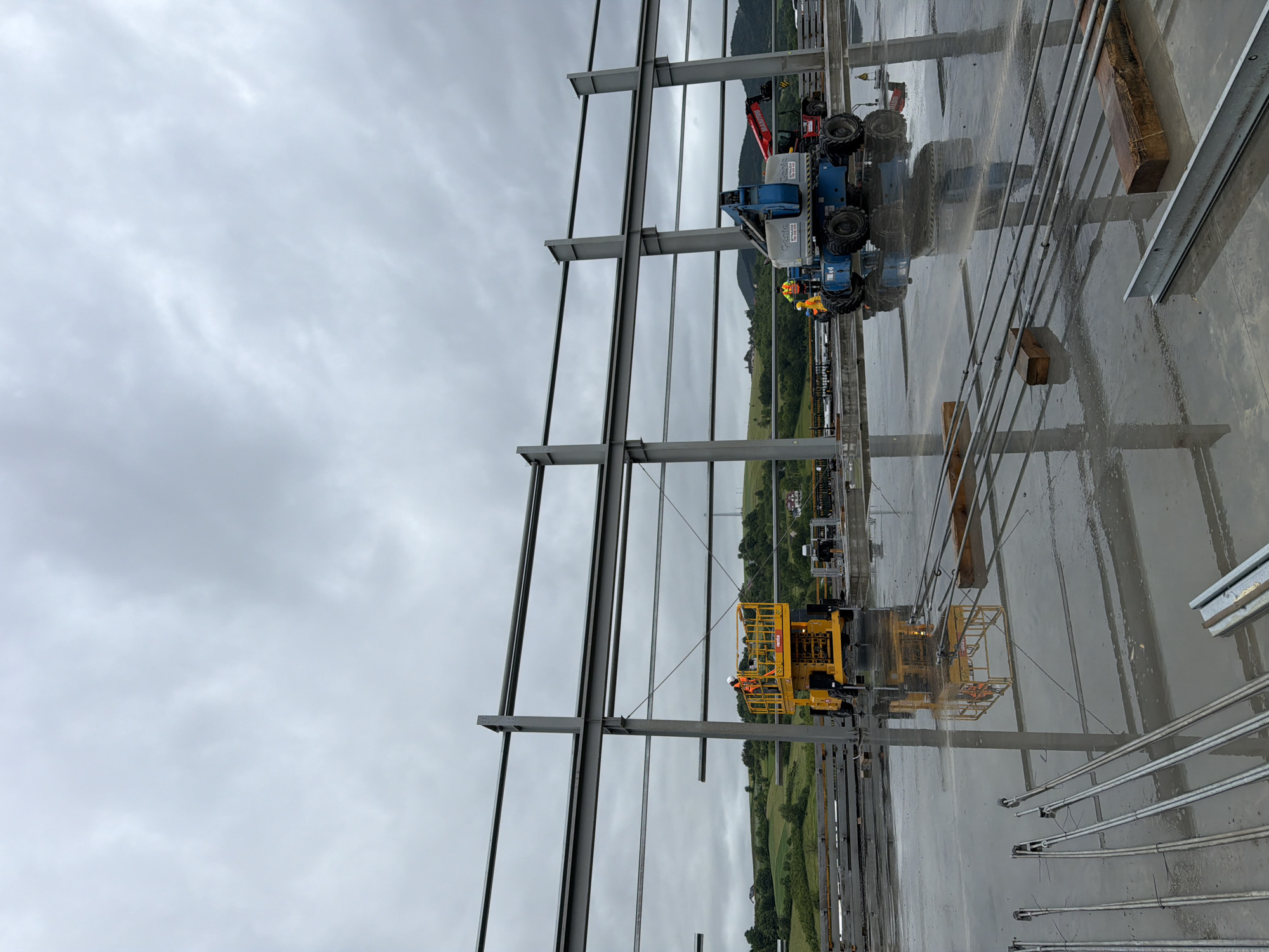 A construction site in Janowice, Poland, shows a large, unfinished steel structure under a cloudy sky. Two aerial work platforms are in use. A yellow platform lift is positioned on the wet, reflective ground near a steel column, with one worker in a high-visibility vest and hard hat in the platform. To the right, a blue articulated boom lift is also on the wet ground, with another worker in a high-visibility vest and hard hat visible in its basket. In the background, rolling green hills and some buildings are visible. The ground is covered in a thin layer of water, reflecting the structure and machinery. Several long, metal rods lie on the ground in the foreground.