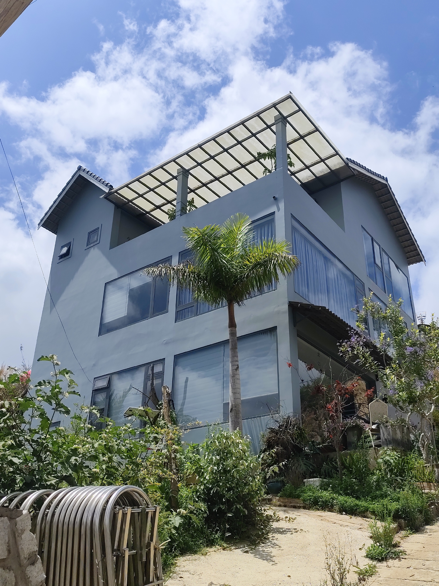 A modern, multi-story building, likely a residence or guesthouse, stands under a bright, partly cloudy sky. The building is painted in a light grey or dusty blue color and features a contemporary design with numerous large, rectangular windows across its facades, many reflecting the sky and surrounding greenery.
A tall palm tree is prominently positioned in front of the building, its fronds reaching past the second floor. The upper level of the building includes a balcony area covered by a translucent, grid-patterned roof structure. The roof of the main structure has a traditional tiled section visible on the left. A small security camera is mounted on the upper left exterior wall.
The foreground and areas surrounding the building are abundant with lush green vegetation, including various bushes, flowering plants with white and purple blossoms, and smaller trees. The ground in the foreground is a sandy or dirt path with sparse weeds. In the lower left corner, a folded, silvery metal barrier or gate, composed of curved segments, is visible. The sky is clear blue with scattered white cumulus clouds, indicating bright daylight and fair weather. There are no people visible, and no specific activity is taking place. No visible text is present. The scene depicts a tranquil, modern dwelling nestled within a verdant setting, consistent with the provided location context of Lạc Dương, Vietnam.