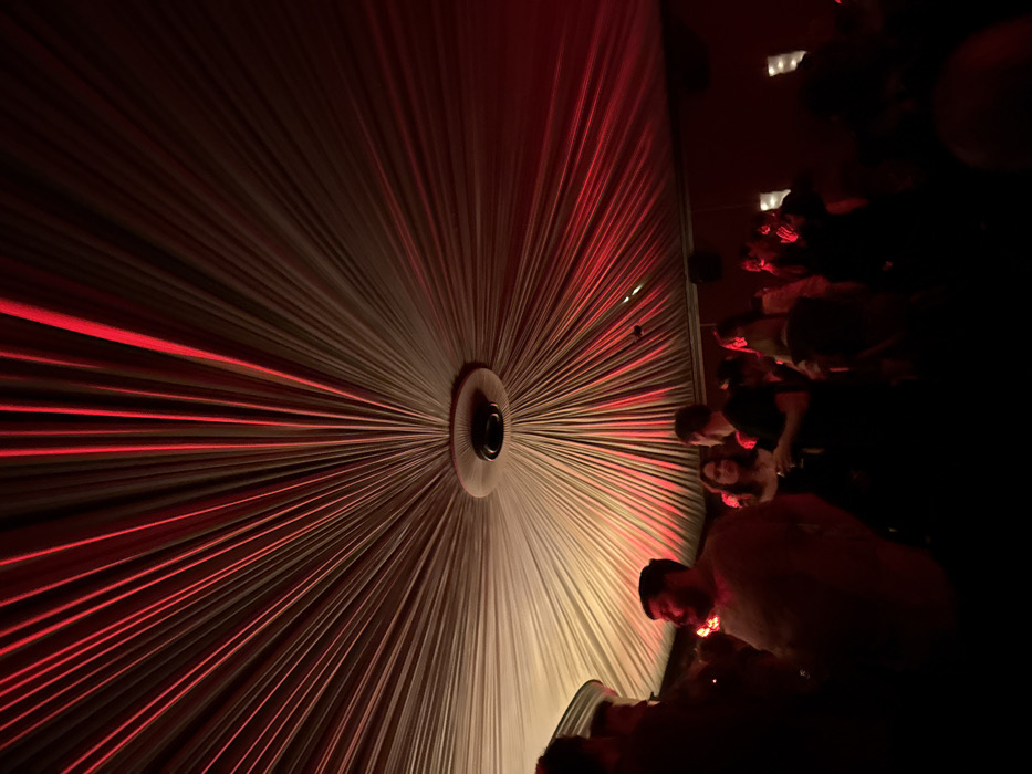 The image depicts an interior scene within a large venue, identifiable as Le Bataclan in Paris, France. The dominant feature is a vast, light-colored fabric ceiling, intricately pleated and draped to radiate from a central circular fixture. Intense red lighting emanates from an unseen source, illuminating the pleated ceiling with strong crimson hues and casting elongated shadows.
Below the illuminated ceiling, a gathering of individuals is present. In the left foreground, a bearded man wearing a light-toned top is partially visible. Towards the mid-ground, a man in a dark shirt holds a glass while appearing to interact with a woman with dark hair standing nearby. Additional figures populate the dimly lit background, largely obscured by shadow and the pervasive red light. Individuals are observed standing and mingling in a social setting, consistent with a nightclub or event space. The central circular ceiling fixture is dark, featuring a recessed element.