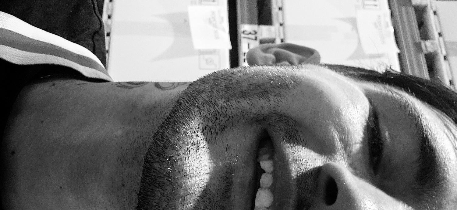 This black and white, close-up image captures a person, likely male, from the neck up, positioned sideways, appearing to be lying down or leaning back. The person's face is illuminated, showing visible stubble or a short beard along their jawline and chin, which glistens under the light. Their mouth is slightly open, revealing white teeth, suggesting a gentle smile or relaxed expression. One ear is visible towards the top of the head.
A faint, circular tattoo pattern is discernible on the skin of the person's neck or upper shoulder. They are wearing a dark garment with a prominent striped trim in white and possibly other colors, though only shades of grey are visible. The background is out of focus but depicts an indoor setting, possibly a storage or retail area, with what appear to be shelves holding white boxes or papers. Visible text in the background includes the numbers "37" and "11" on labels. The lighting creates strong highlights on the person's facial hair, forehead, and teeth, adding depth and texture to the monochrome portrait. The overall impression is one of calm or quiet amusement. The scene is set in Plovdiv, Bulgaria.