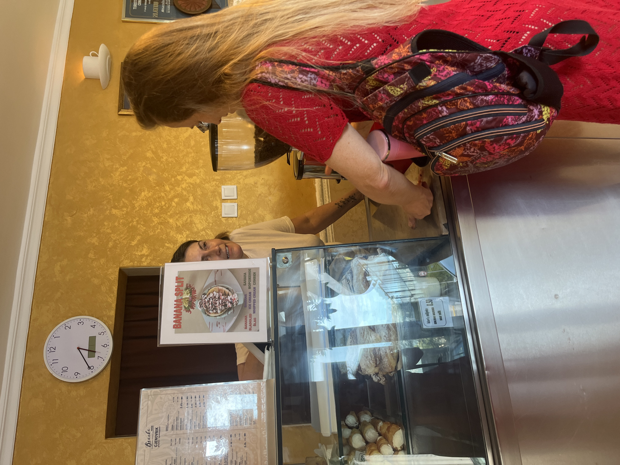In Budva, Montenegro, a woman in a red dress with a floral backpack stands at a counter in what appears to be an ice cream parlor or cafe, facing a glass display case. She is interacting with a female employee who is leaning over the counter, looking at her. Behind the employee, a framed menu and a "Banana Split" menu are visible on the wall. To the left, a large clock shows approximately 4:18. A coffee grinder is visible behind the employee. The display case contains various pastries and items, with a small sign indicating "kiselo mlijeko" for 0.50. The environment features a textured yellow wall and metallic surfaces of the counter.