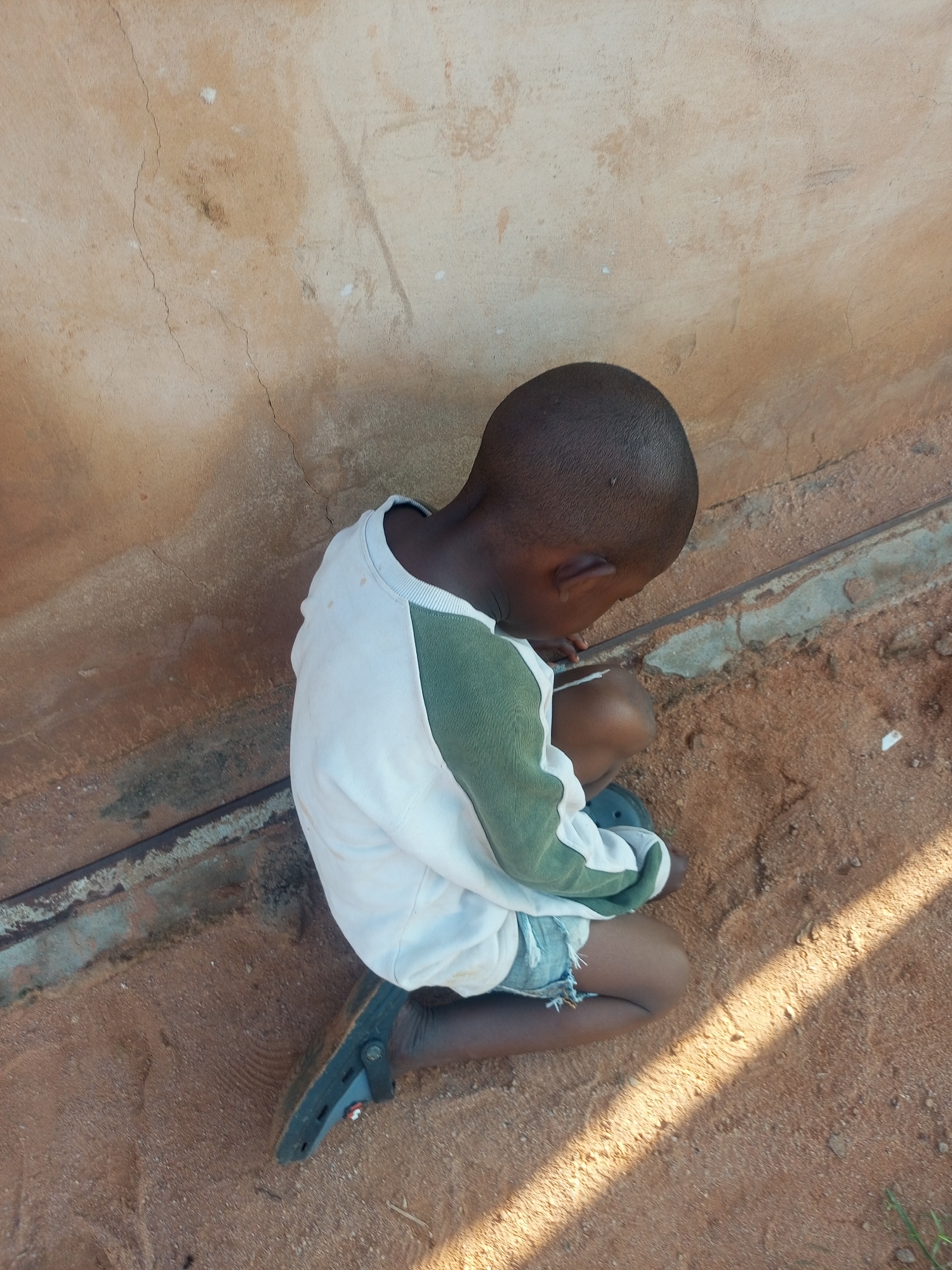 A young boy is sitting on sandy ground next to a textured, light brown wall with visible cracks. He is wearing a white long-sleeved shirt with green panels on the sleeves and shoulders, and denim shorts. His head is bowed, and his gaze is directed downwards. A dark-colored shoe is on his right foot. The lighting suggests it is daytime, with sunlight casting shadows on the ground.