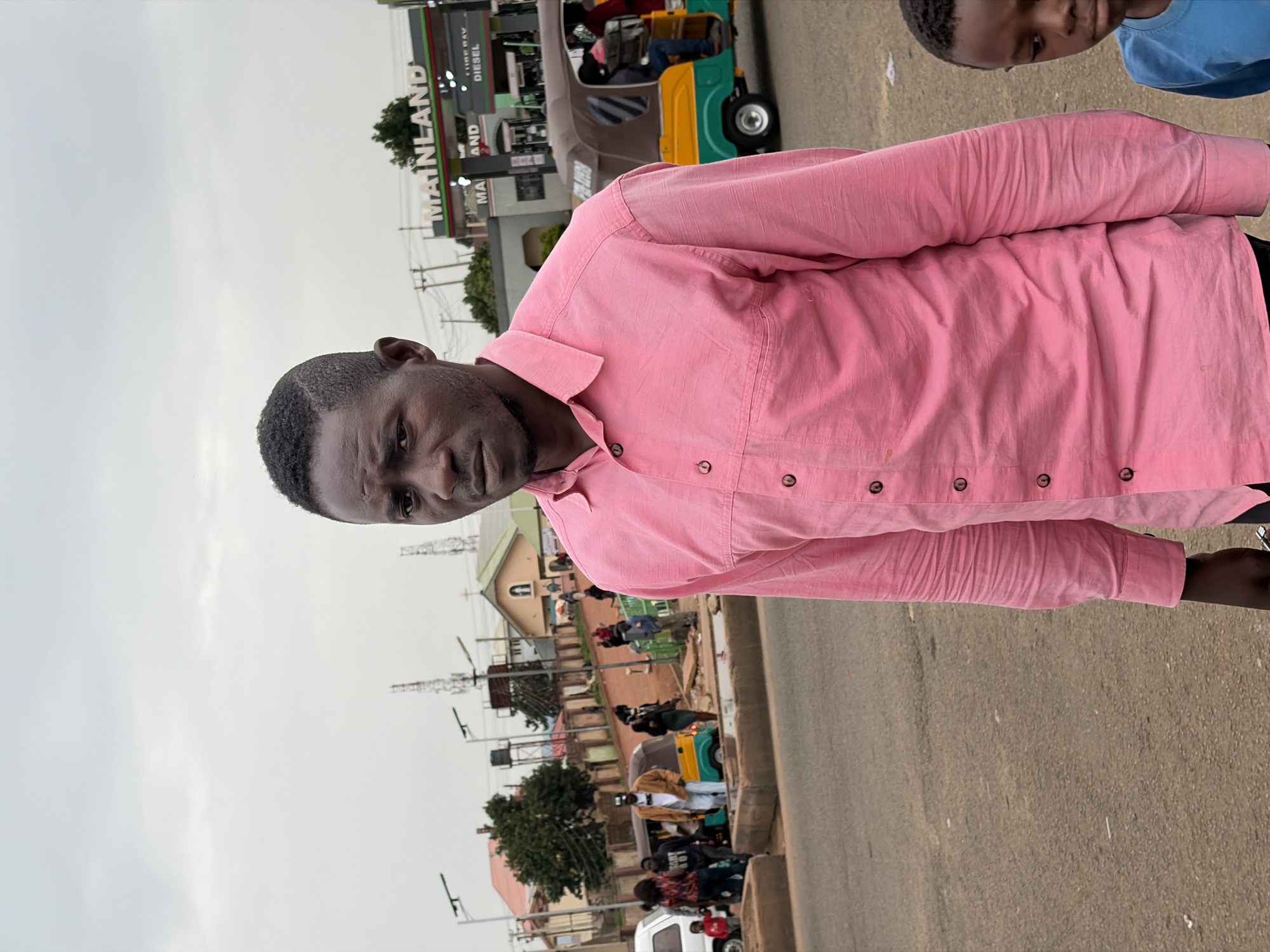 A medium shot captures a man in a pink collared shirt standing on a paved road in Gura Top, Nigeria. The man is positioned in the foreground, facing slightly to his right with a neutral expression. Behind him, the scene unfolds with a gas station branded "MAINLAND" on the right side of the frame, featuring pumps and signage. A three-wheeled auto-rickshaw is visible, with passengers inside. Further in the background, several buildings with pitched roofs are present, including what appears to be a church with a cross. Various people are scattered throughout the scene, some walking, others near vehicles. The environment suggests a bustling urban or semi-urban area with overcast skies. A young boy in a blue shirt stands to the right of the main subject, partially visible.