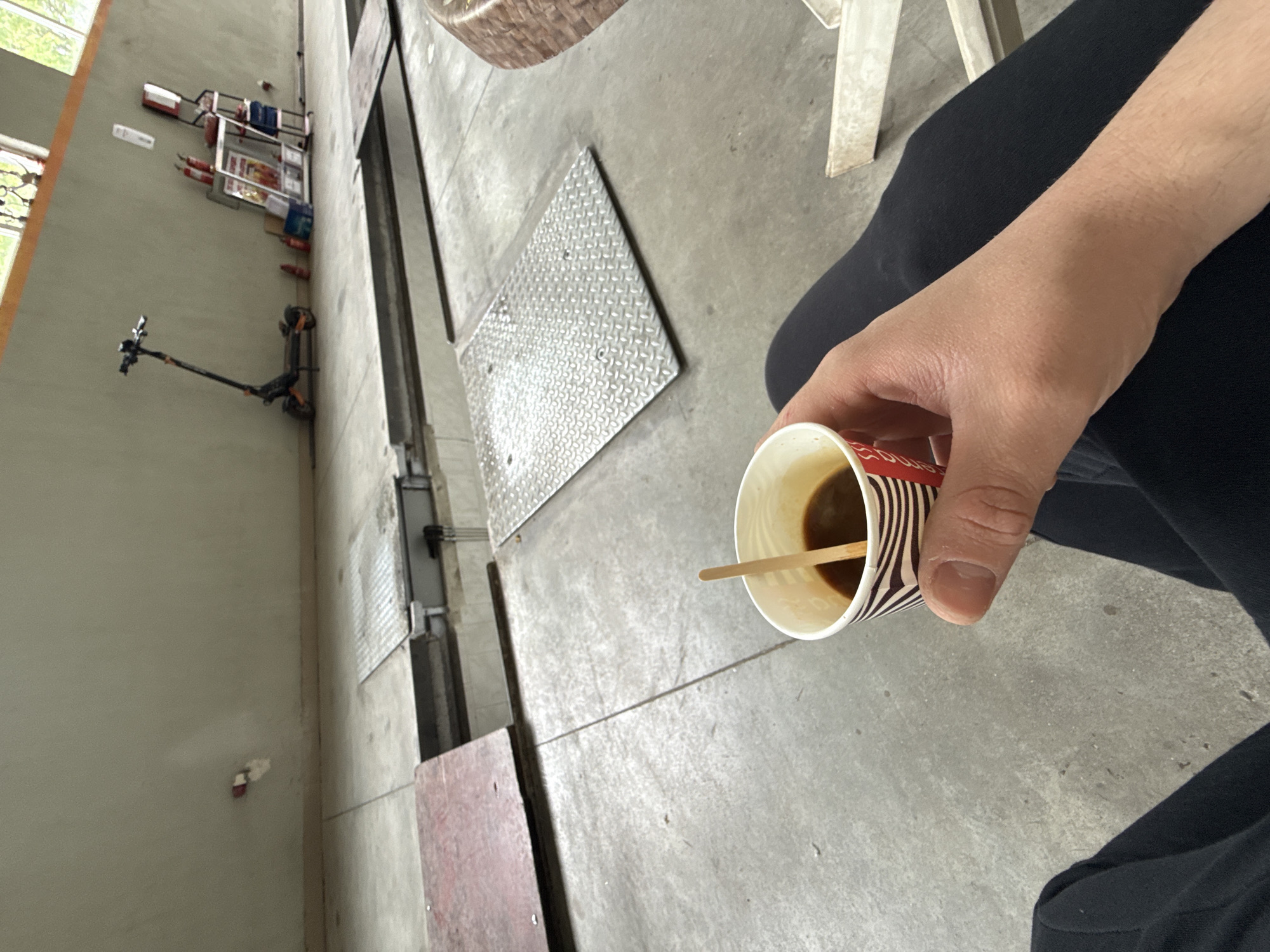 A first-person perspective shot from an industrial setting in Stara Zagora, Bulgaria. The primary focus is a hand holding a disposable coffee cup with a wooden stirrer inside. The background includes elements like a scooter, a tool storage rack mounted on the wall, a metal plate, industrial flooring, and the corner of a white chair, suggesting the image was taken in a warehouse or workshop environment. The person is wearing black pants.