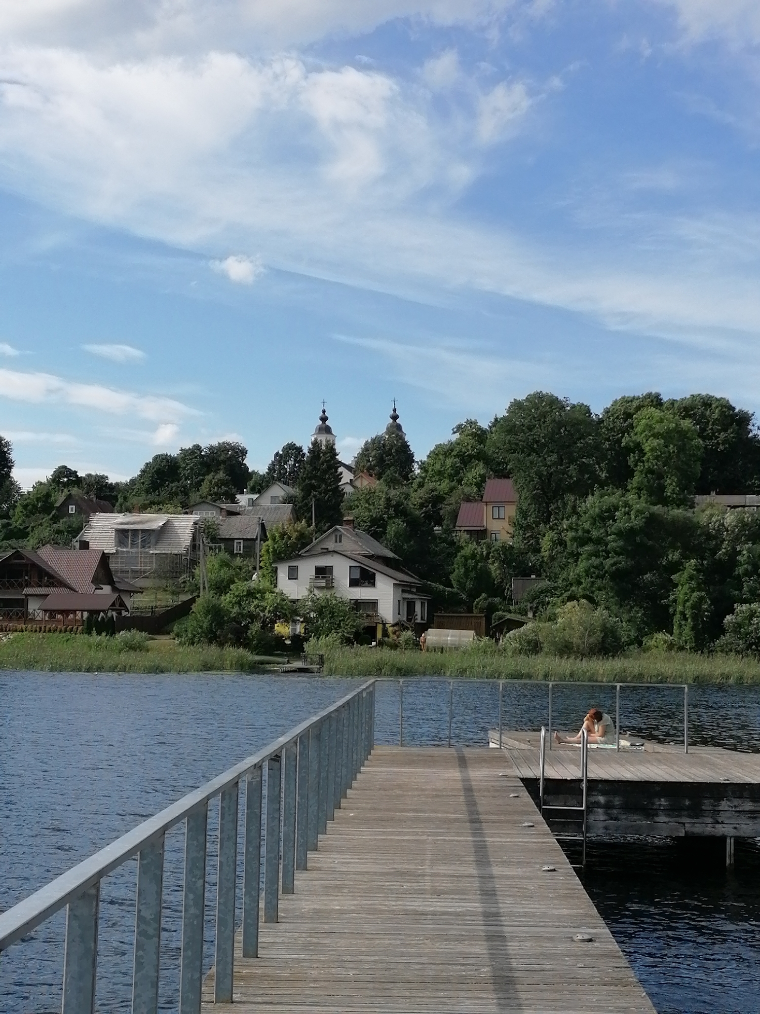 The image depicts a scenic view of a lake in Zarasai, Lithuania, on a clear day with scattered clouds. In the foreground, a wooden pier with a metal railing extends into the blue water. A lone person, with reddish hair, is sitting on the end of the pier, facing away from the camera. The person is wearing a light-colored top and appears to be relaxing or sunbathing.
Beyond the lake, a cluster of houses and trees lines a gentle slope. Several houses are visible, with varying architectural styles. One house is under construction, with scaffolding around its roof. A church steeple with a cross is prominent among the buildings. The trees are lush and green, suggesting a summer or late spring setting. The water of the lake is calm, reflecting the blue sky and clouds. Reeds and tall grasses grow along the shoreline. The overall impression is one of tranquility and a pleasant summer day by the water.