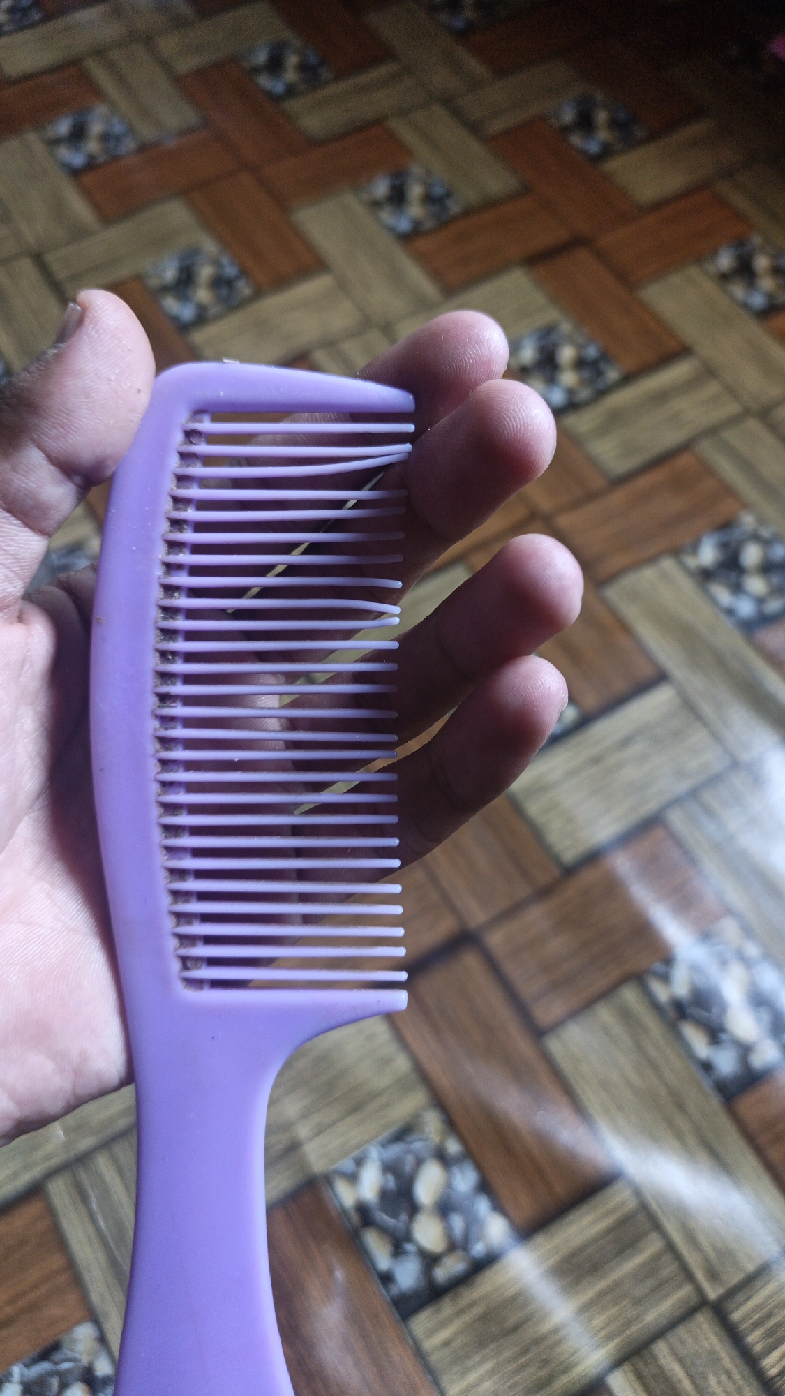 A person's left hand holds a purple comb with several strands of yellow hay or grass caught between its teeth. The comb is positioned vertically, with the teeth facing upwards. The background is a tiled floor with a wood-like pattern, featuring alternating brown and lighter wood-colored rectangles arranged in a diagonal herringbone pattern. Small, speckled stone-like insets are visible within some of the rectangles. The lighting suggests an indoor environment, possibly a home, and the overall scene is focused on the comb and the hand. There is no identifiable location in Sungai Rasau, Indonesia, present in the image.
